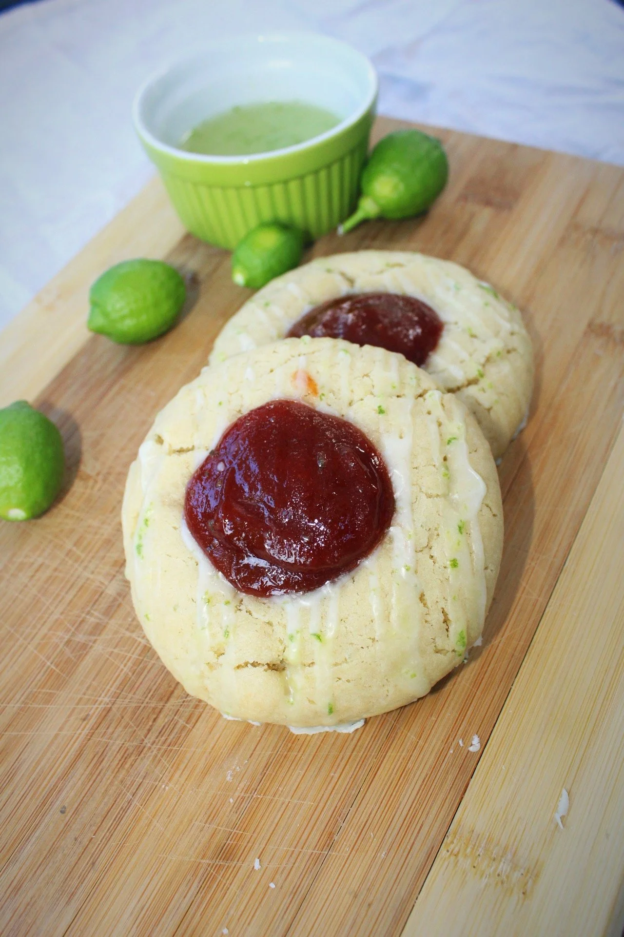 Two cookies with jam and icing on a wooden board, along with limes and a bowl of green juice.