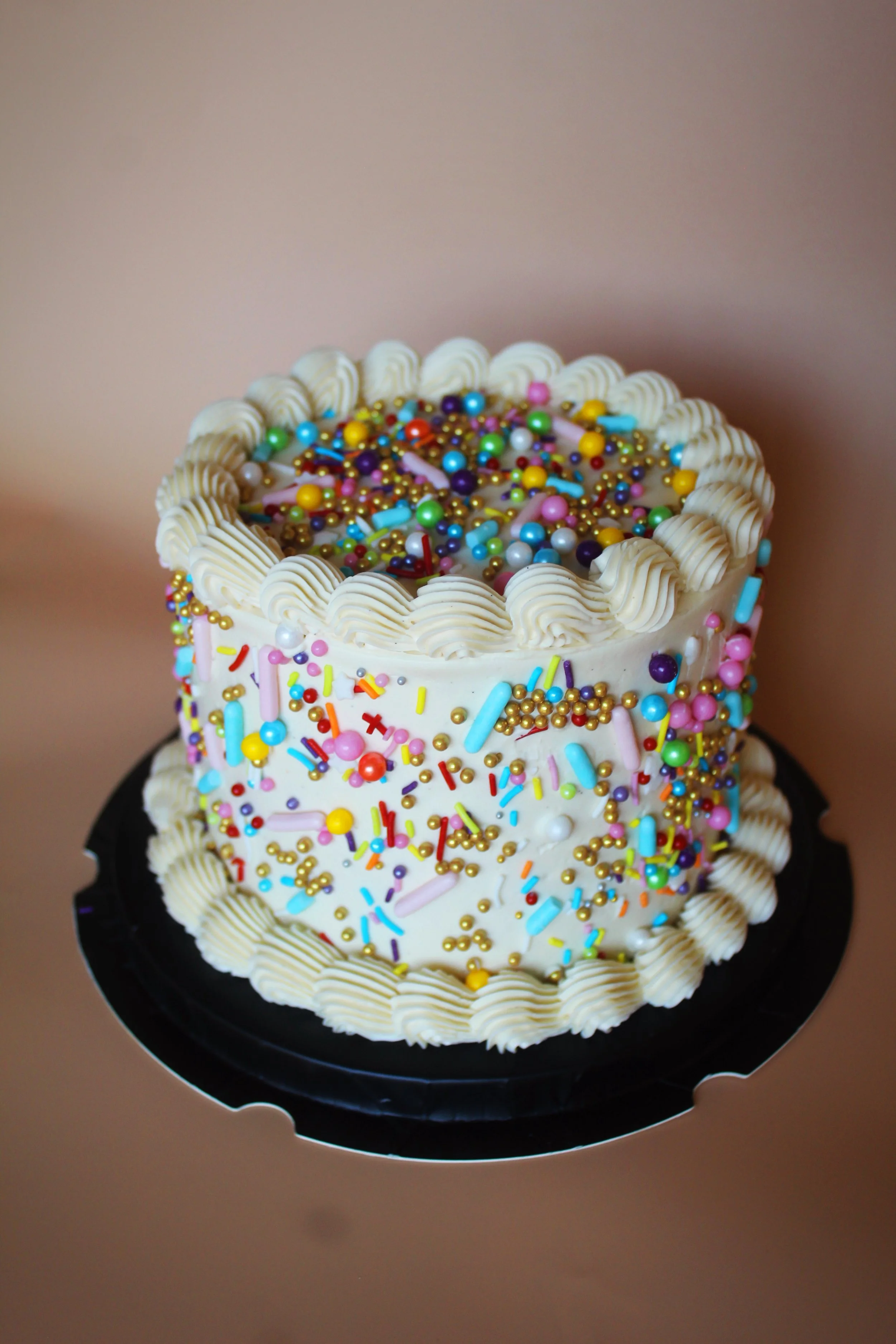 A multi-layered birthday cake decorated with white frosting, colorful sprinkles, and small edible beads, with piping around the edges, placed on a black cake stand against a beige background.