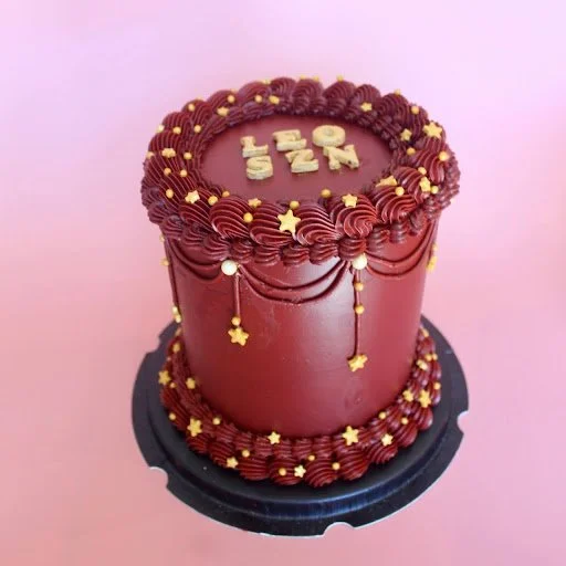 A tall birthday cake with red frosting, decorated with ruffled borders and gold star sprinkles, featuring the number 5 and letters 'L' and 'O' on top, placed on a black cake stand against a pink background.