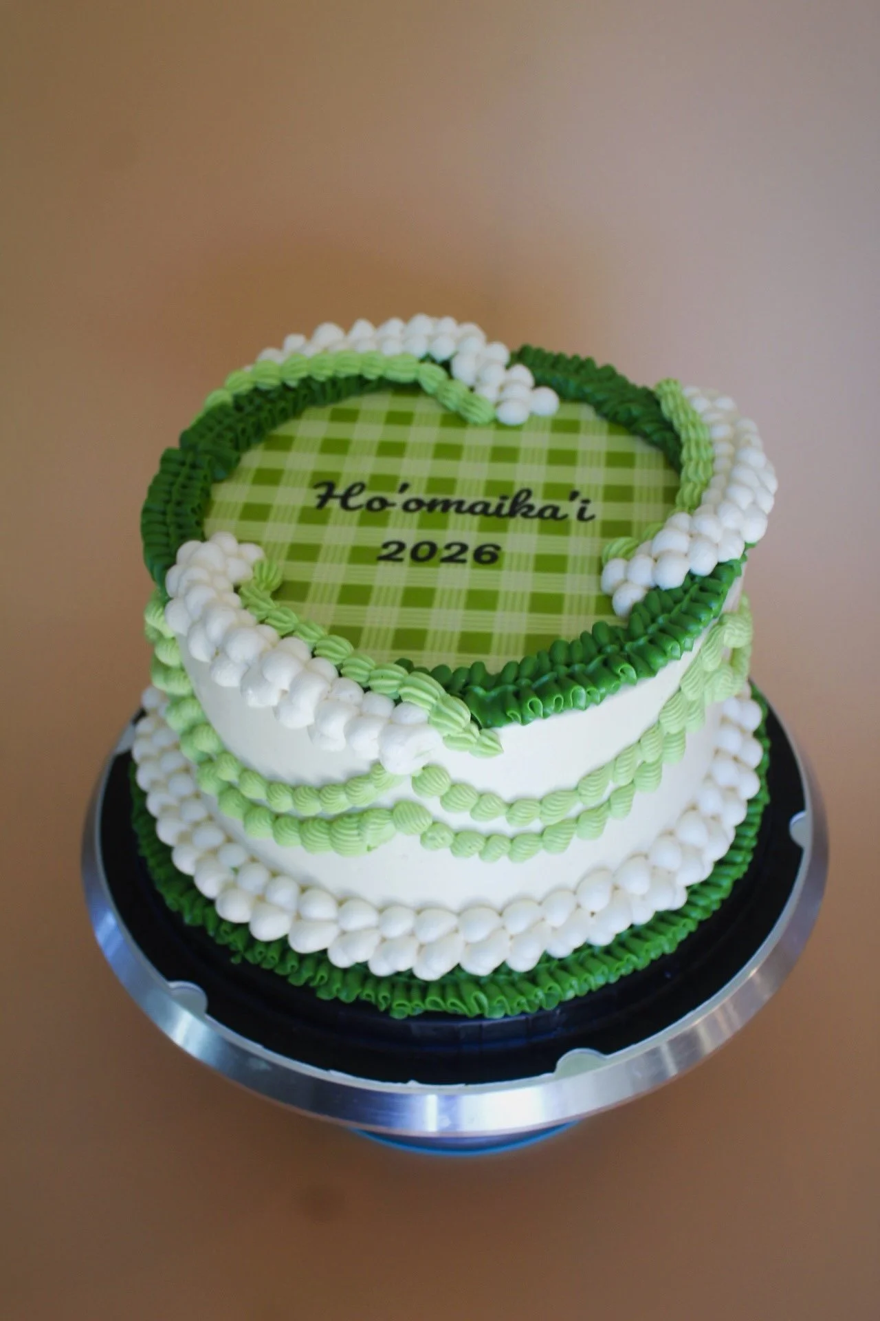 Two-tiered cake decorated with green and white icing, with a green checkered top, and the message 'Ho'omaika'i 2026' written on the top tier.