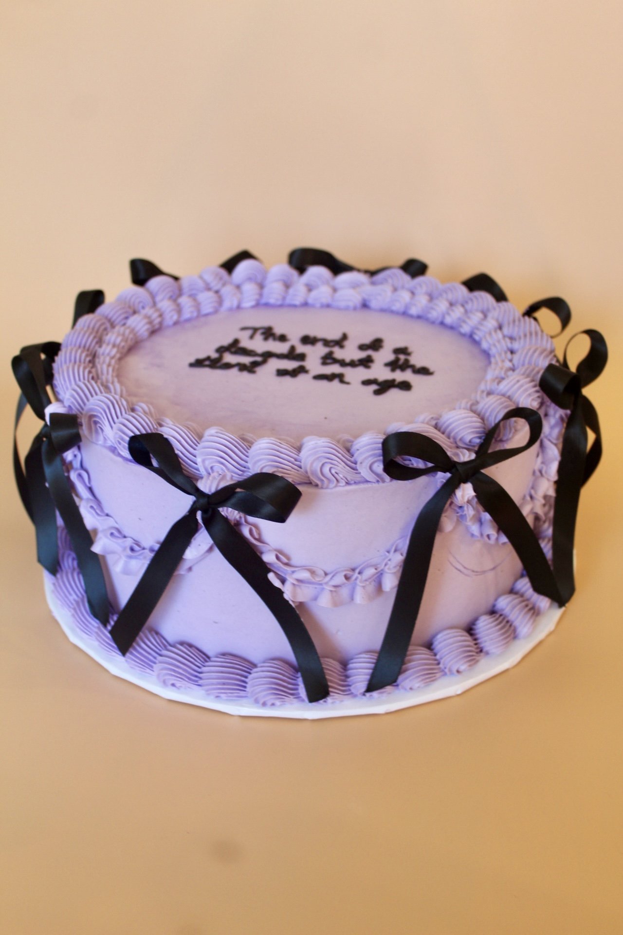 A round birthday cake decorated with purple frosting and black ribbons, with a message written in black icing on top that says, 'The end of an era and a new beginning'.