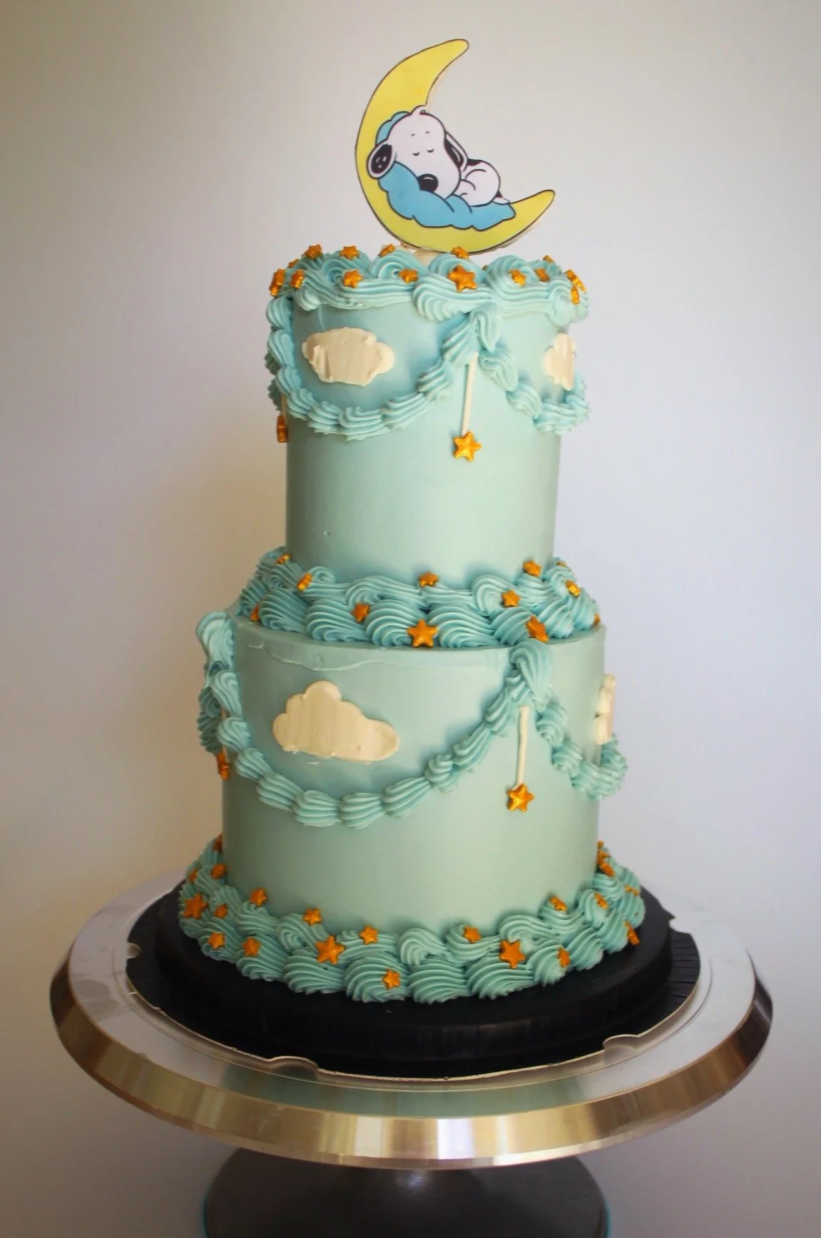 Three-tiered light blue cake decorated with cloud patterns and orange stars. Topped with a Snoopy figurine sleeping on a crescent moon with a star.