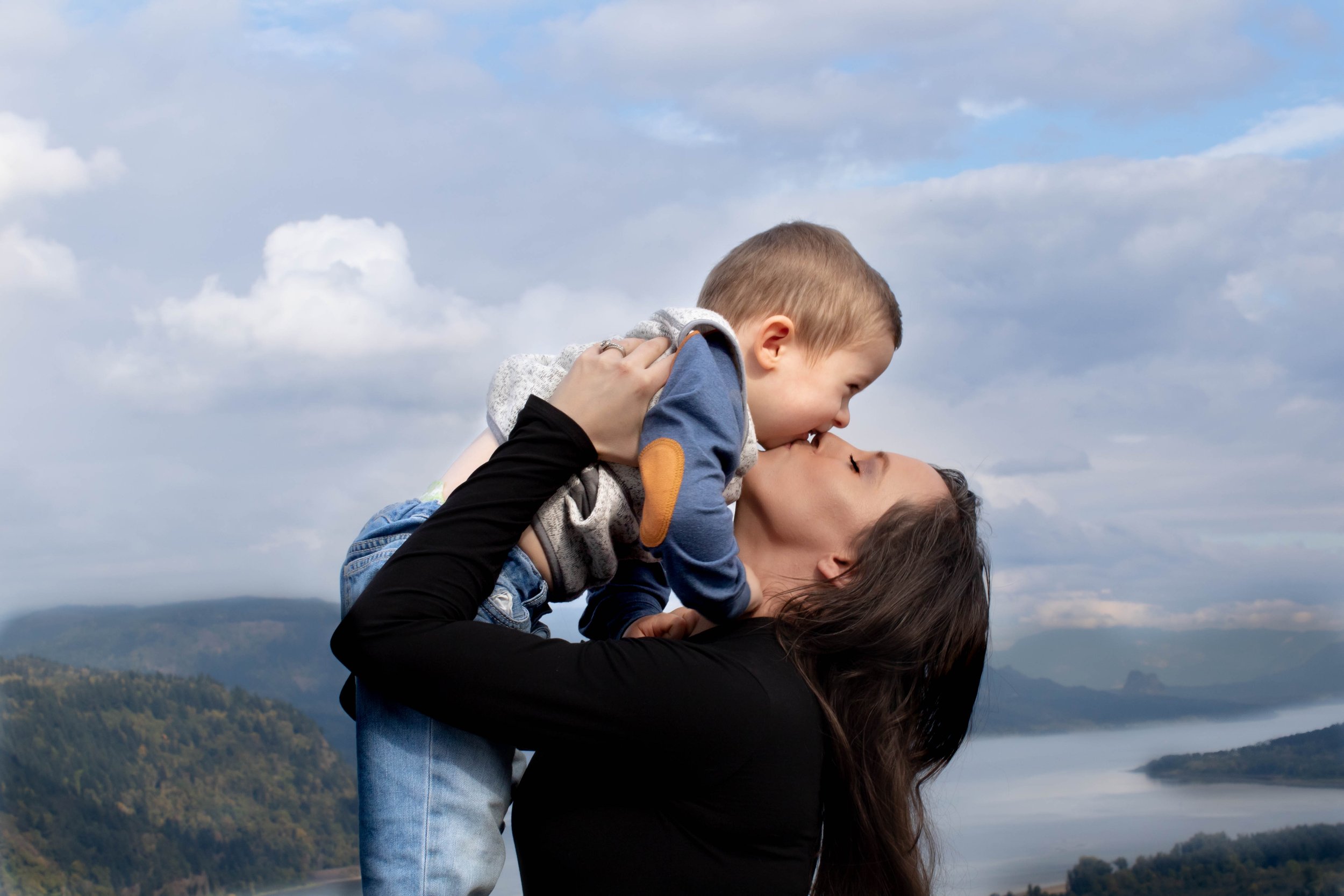 A woman lifts a young boy into the air outdoors, both smiling and looking at each other, with scenic mountains and a partly cloudy sky in the background.