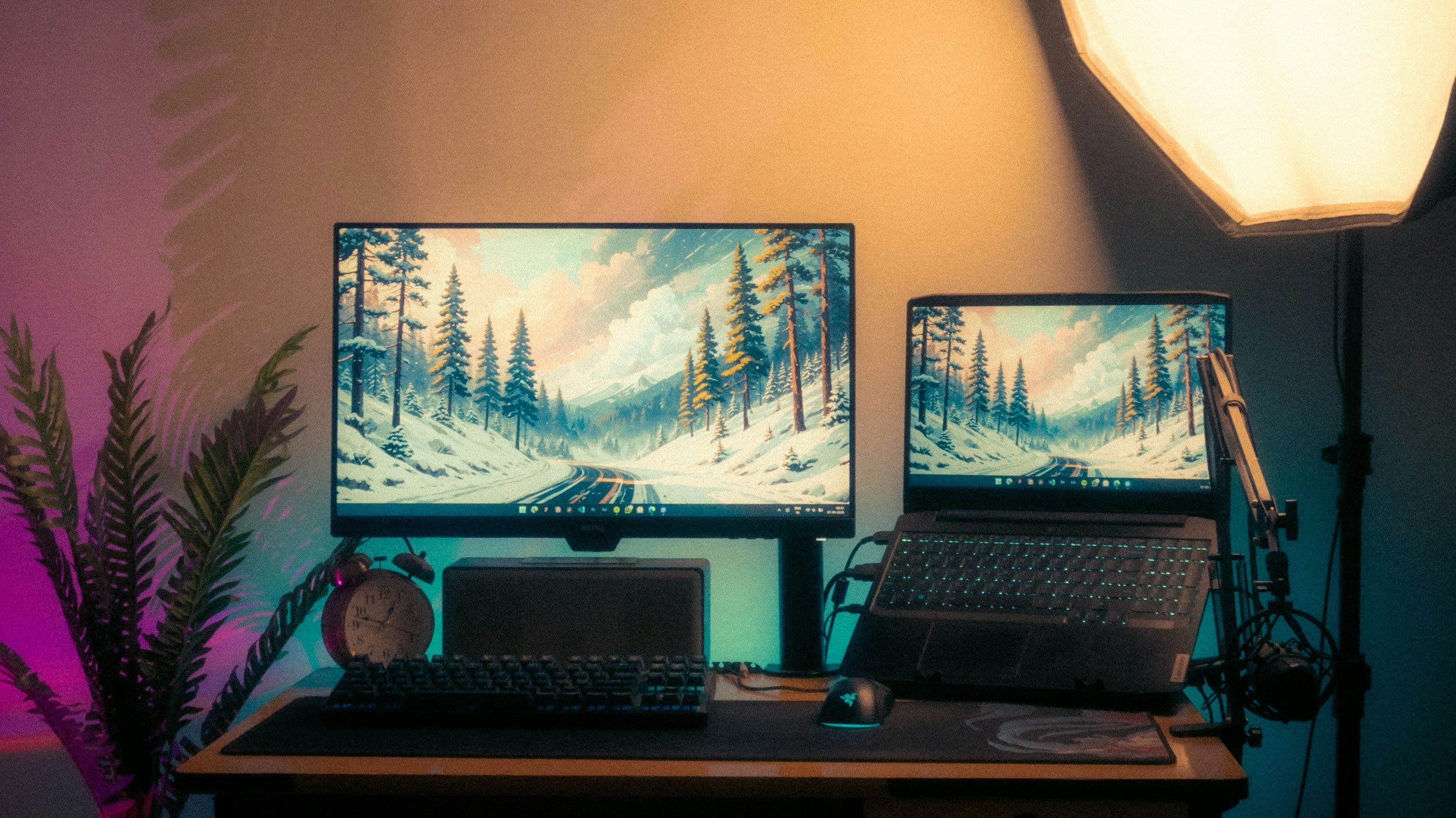A computer desk setup with two monitors displaying a snowy mountain and forest scene, surrounded by a keyboard, mouse, speakers, a clock, a potted plant, and a soft overhead light.