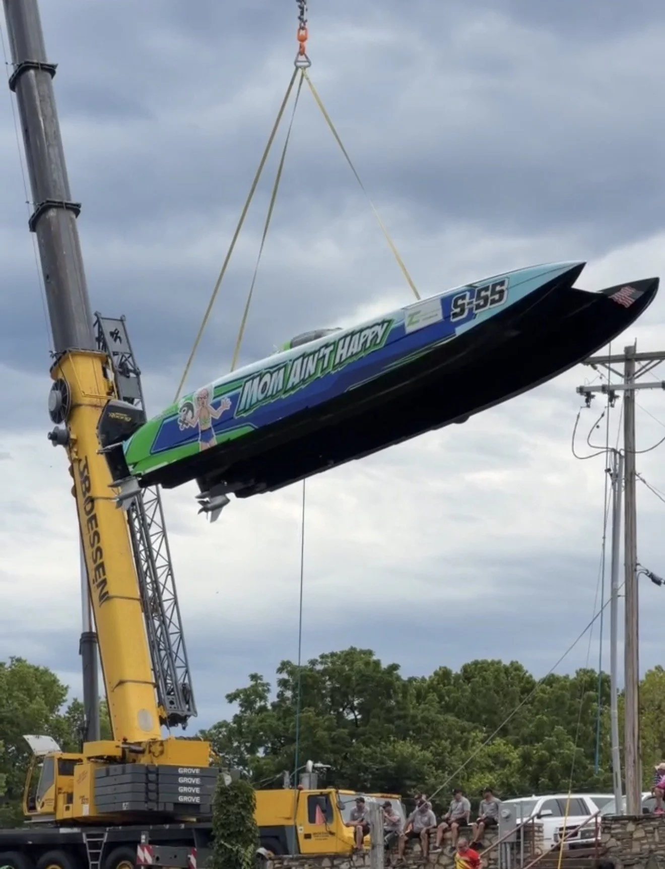 A crane lifting a boat labeled 'Mount Happy' with a cartoon character, in a parking lot with trees and a cloudy sky in the background.