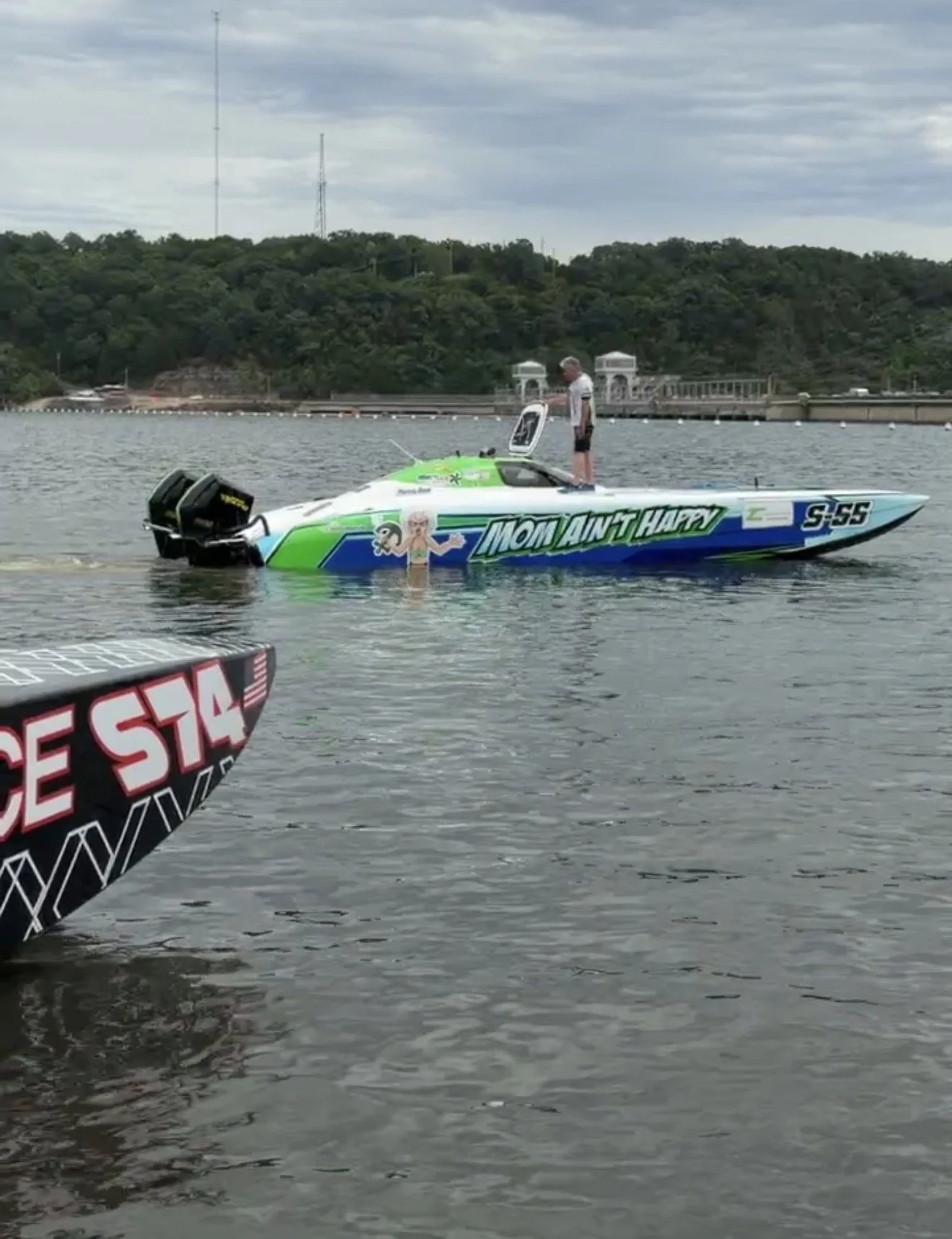 A person standing in a small green and white speedboat on a body of water, with another vessel partially visible in the foreground. The speedboat has a decal that reads 'Mom Ain't Happy' and an illustration of a woman. The background features a hill 
