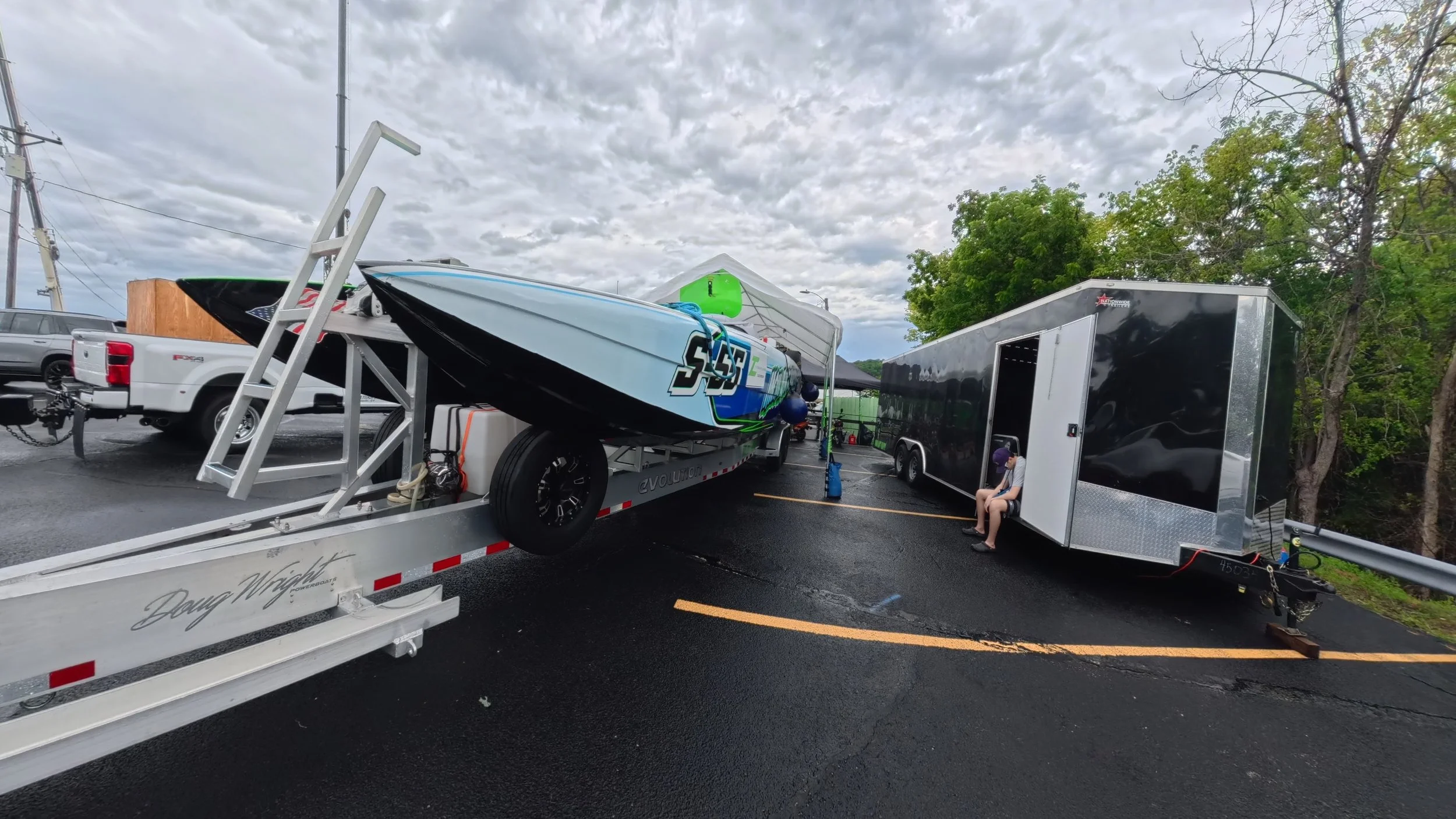 A parking lot with a racing boat on a trailer, a black enclosed trailer, and a person sitting on the black trailer, under a cloudy sky.