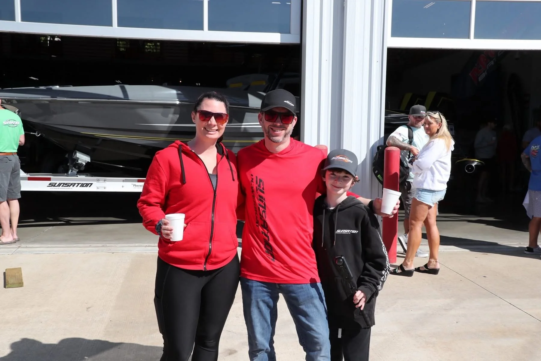 Group of three people smiling and holding cups in front of a boat garage, with other people in the background at a boat show or marina.