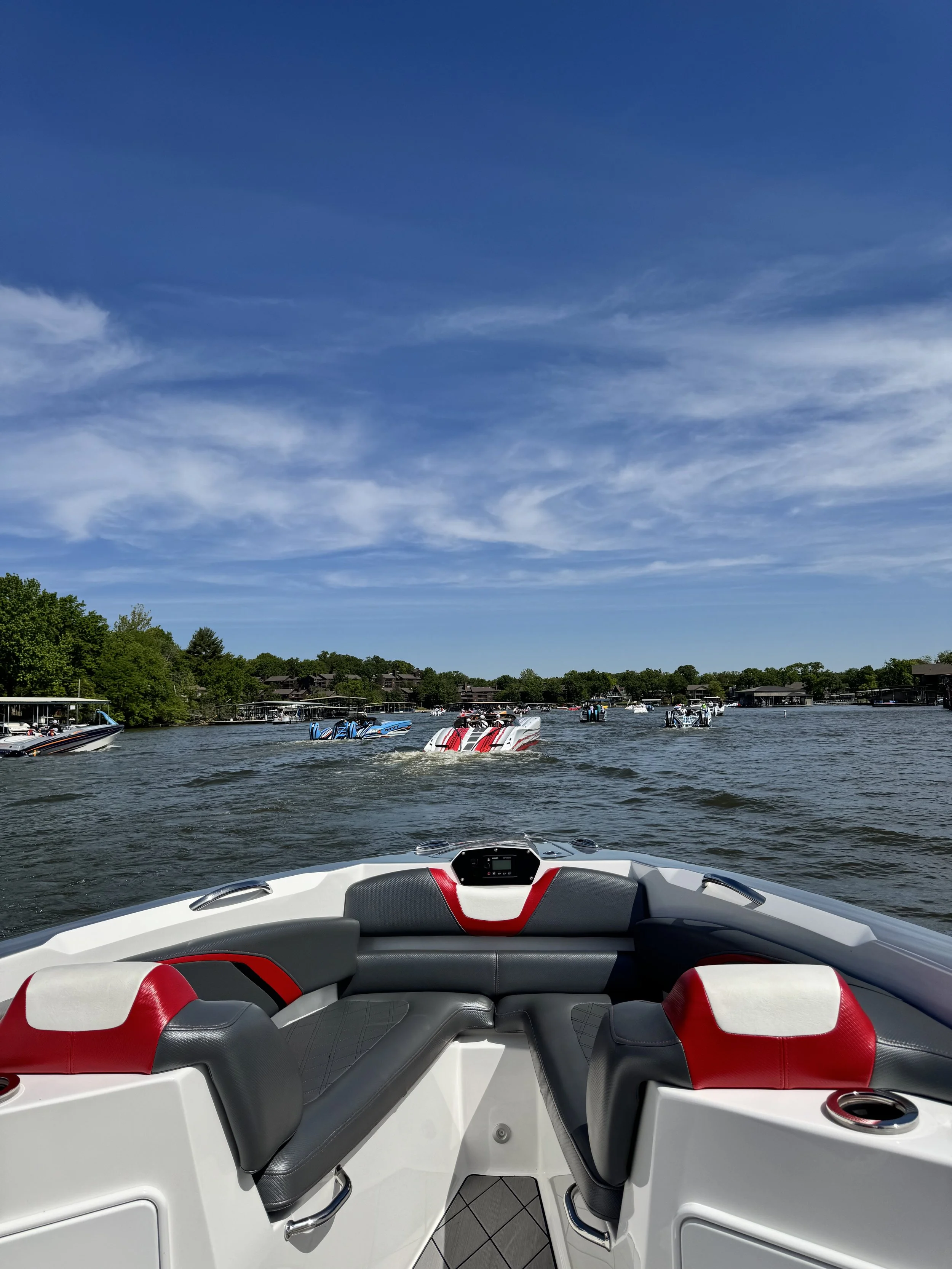 View from the interior of a speedboat showing the dashboard and seating, with several other boats on the water and houses along the shoreline under a blue sky with wispy clouds.