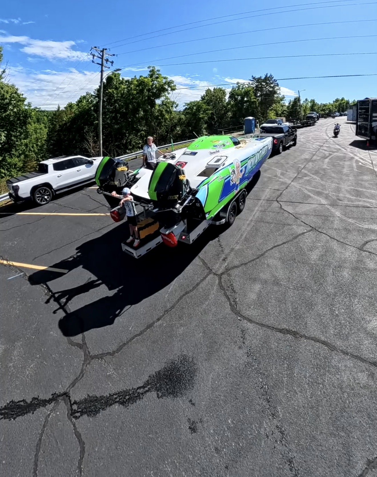 A race boat with a green and white exterior on a trailer in a parking lot, with a man standing beside it and another person nearby. Several vehicles, trees, and a blue sky with some clouds are visible in the background.