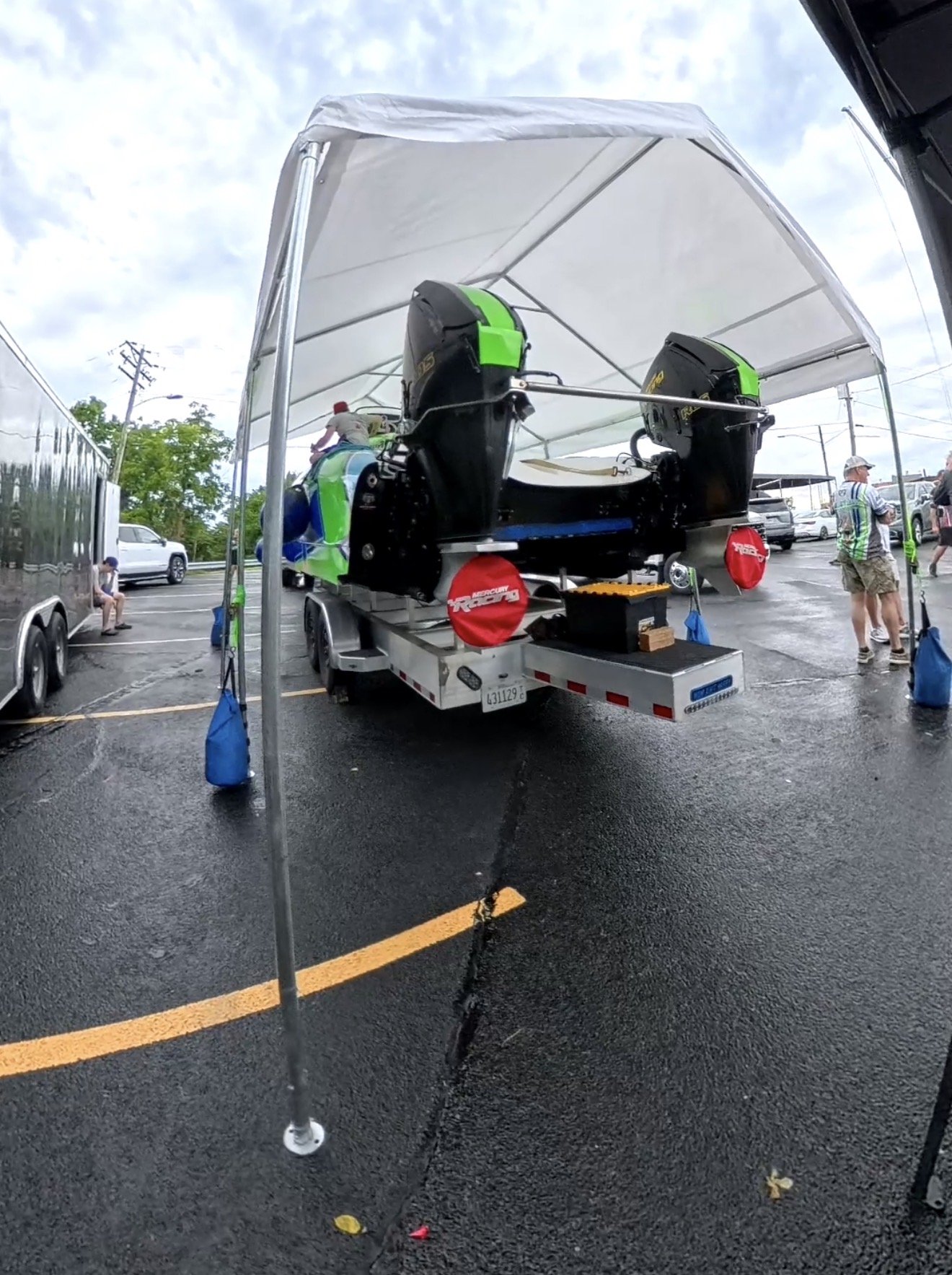 A boat with two outboard motors on a trailer, covered by a white canopy, is parked under a canopy in a parking lot. People are standing and sitting nearby, with cars visible in the background. The parking lot has wet pavement and a large crack.