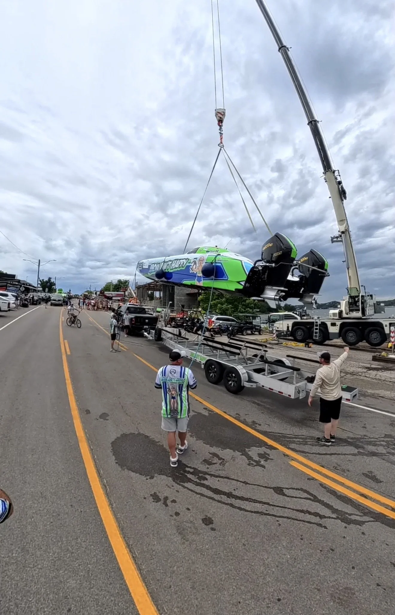 A boat being hoisted into the sky by a crane at a busy parking lot or event. People are watching and taking pictures, with many parked cars and a cloudy sky in the background.
