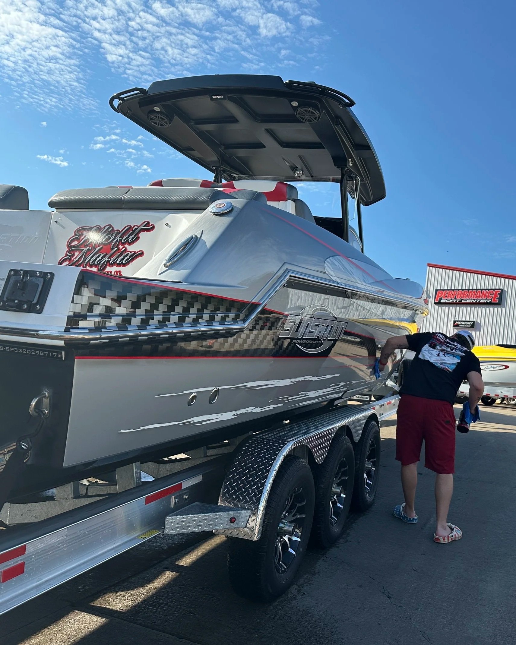 A speedboat on a trailer at a performance boat center, with a person in shorts and a t-shirt standing nearby. The boat has the words "Hotnet Mafia" on its side and is outdoors under a clear blue sky.
