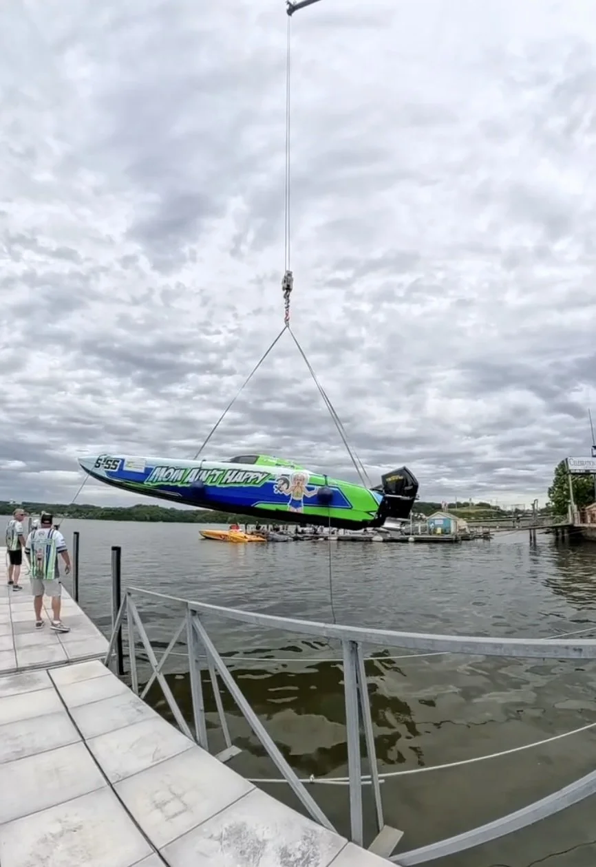 A boat with colorful graphics and the words 'Mom and Happy' is being lifted out of the water by a crane at a dock. There are two people walking on the dock and a yellow kayak in the background.