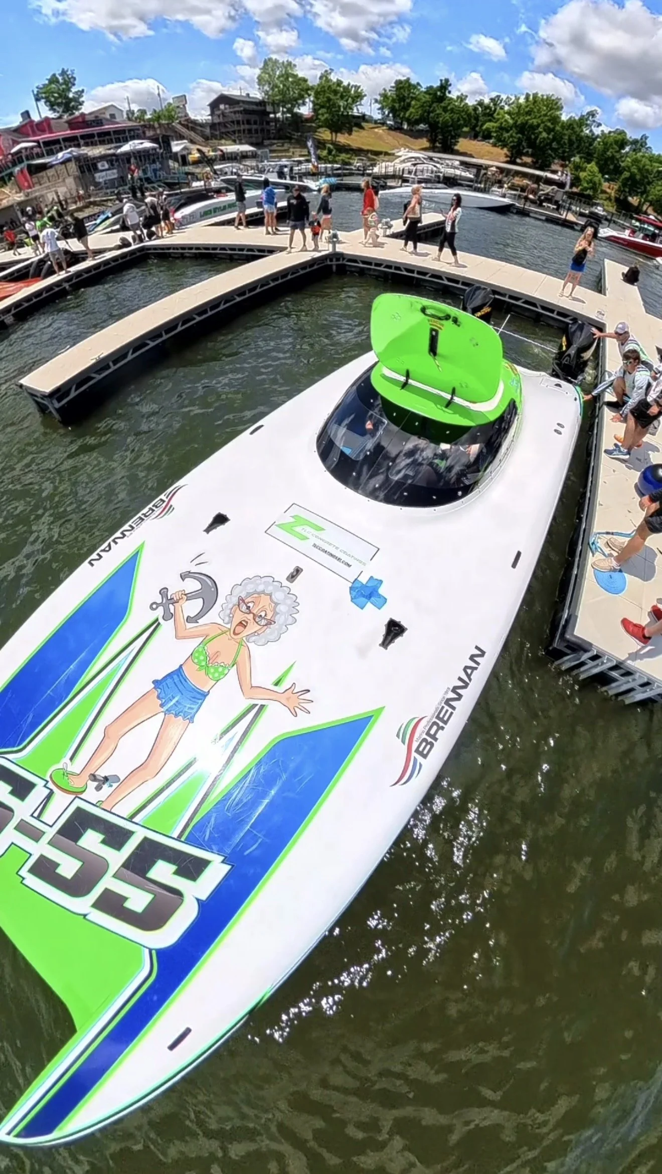 A large speedboat with bright graphics and a cartoon character on its surface, docked at a marina filled with boats and people, with a wooden pier and buildings in the background under a partly cloudy sky.