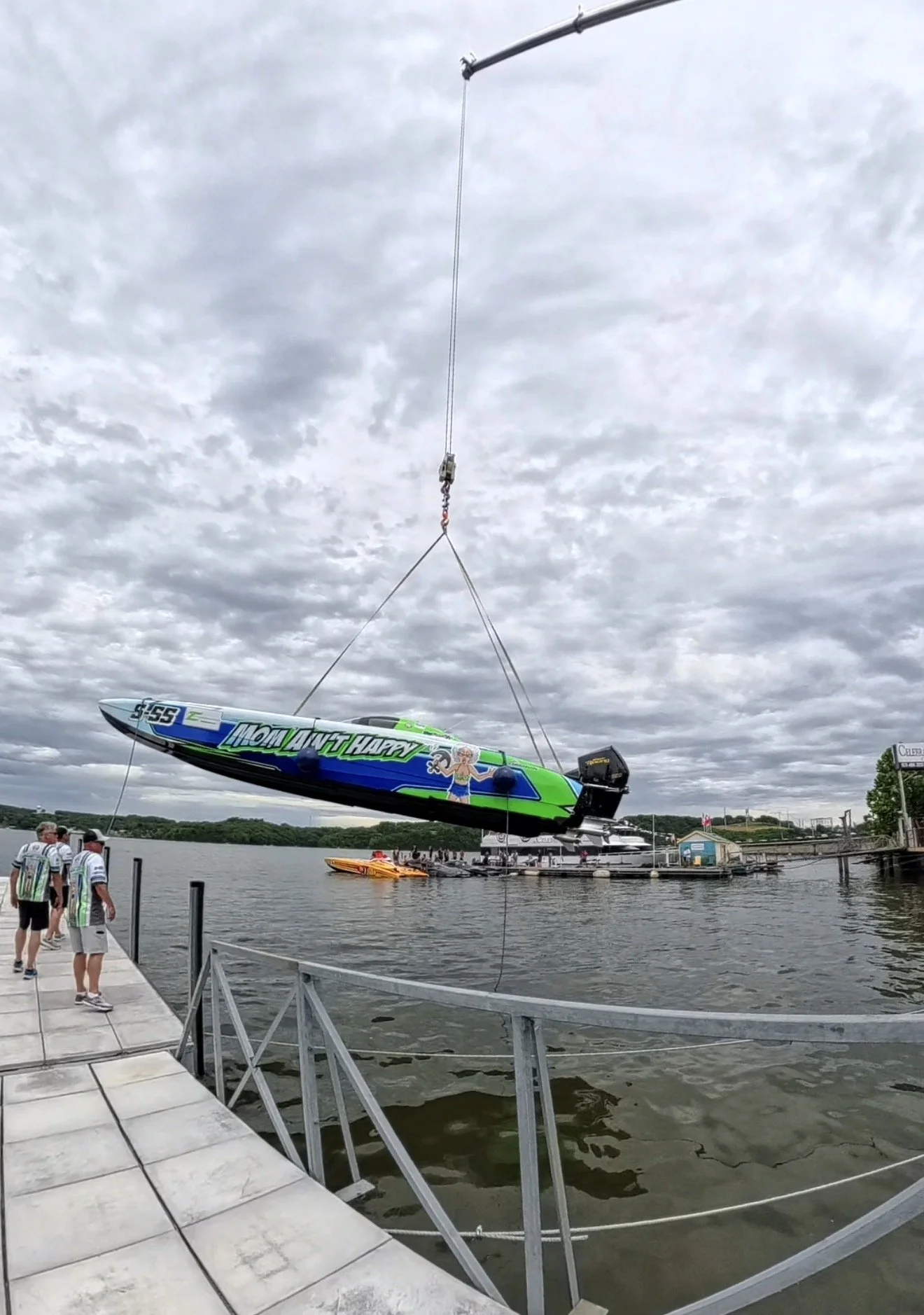 A large motorboat with colorful graphics and characters, hanging from a crane over a body of water at a marina, with people walking on the dock and a cloudy sky above.