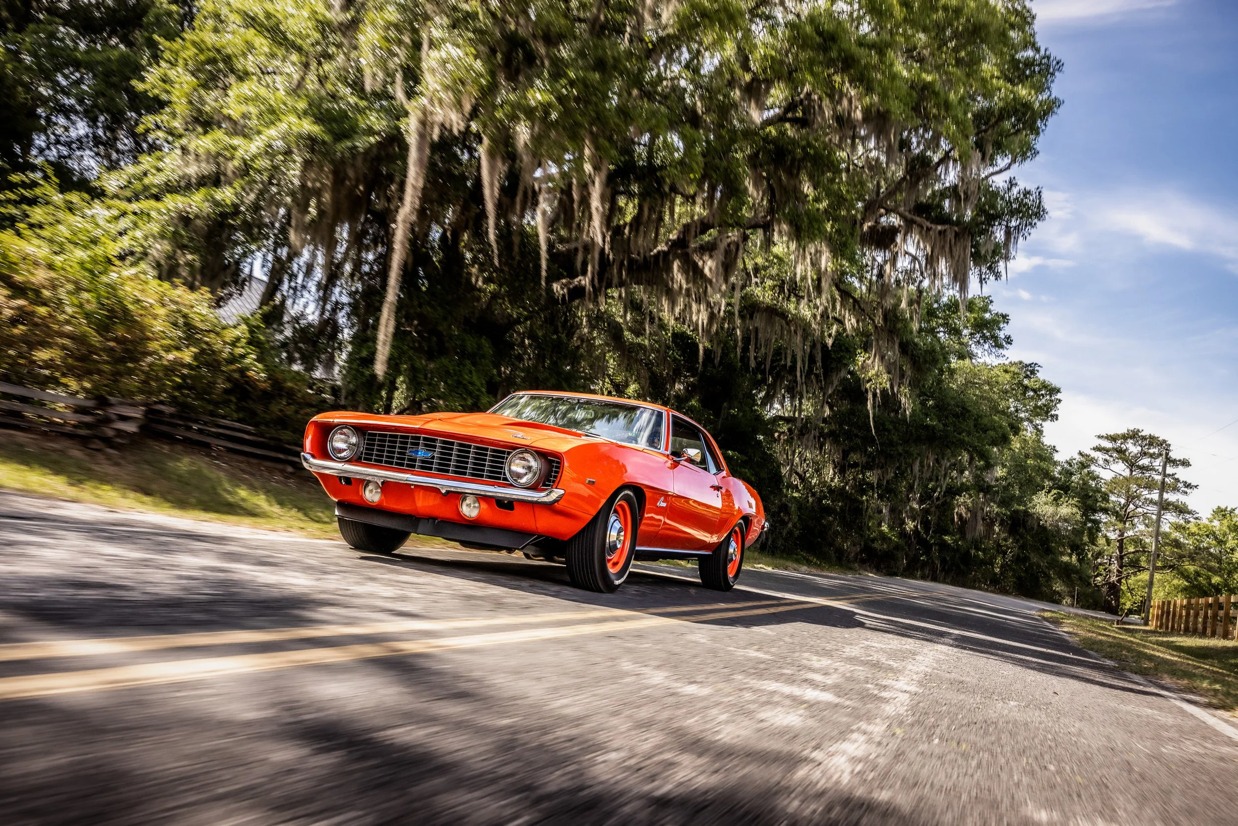 A vintage orange Chevrolet Camaro driving on a rural road beside lush green trees with Spanish moss hanging from the branches.