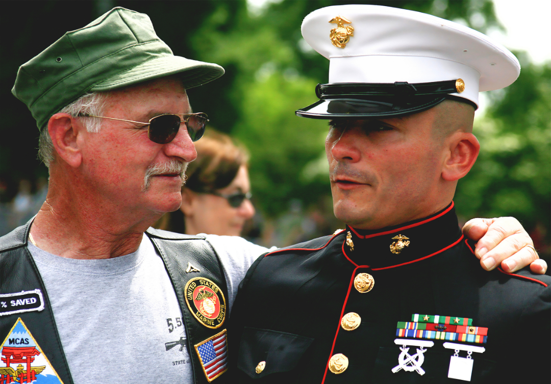 A veteran man in a green cap and sunglasses is comforting a Marine in dress uniform, with a background of trees and blurred people.