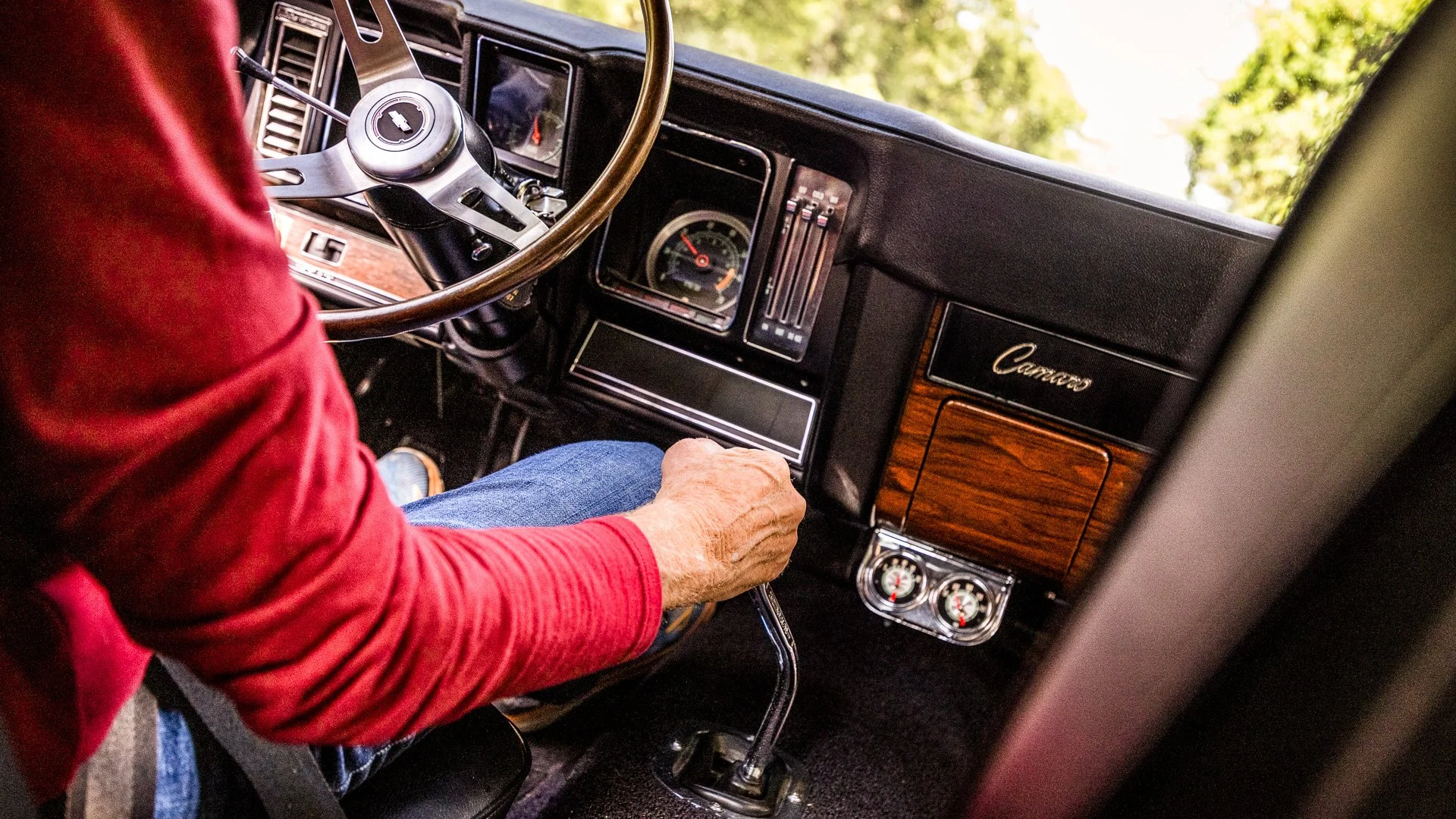 Interior view of a vintage Chevrolet Camaro car, showing the dashboard, steering wheel, and a person in a red shirt driving.