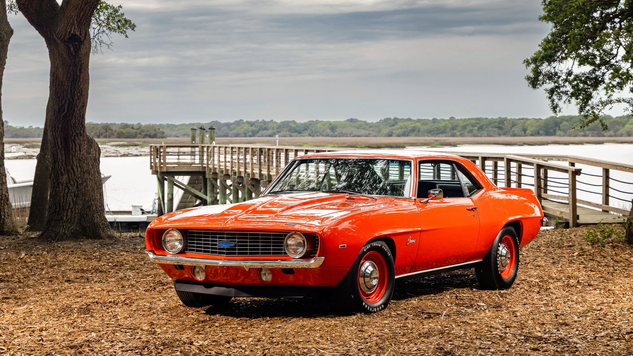 A bright orange vintage Chevrolet Corvair car parked on a dirt area under trees near a body of water, with a wooden dock in the background, during overcast weather.