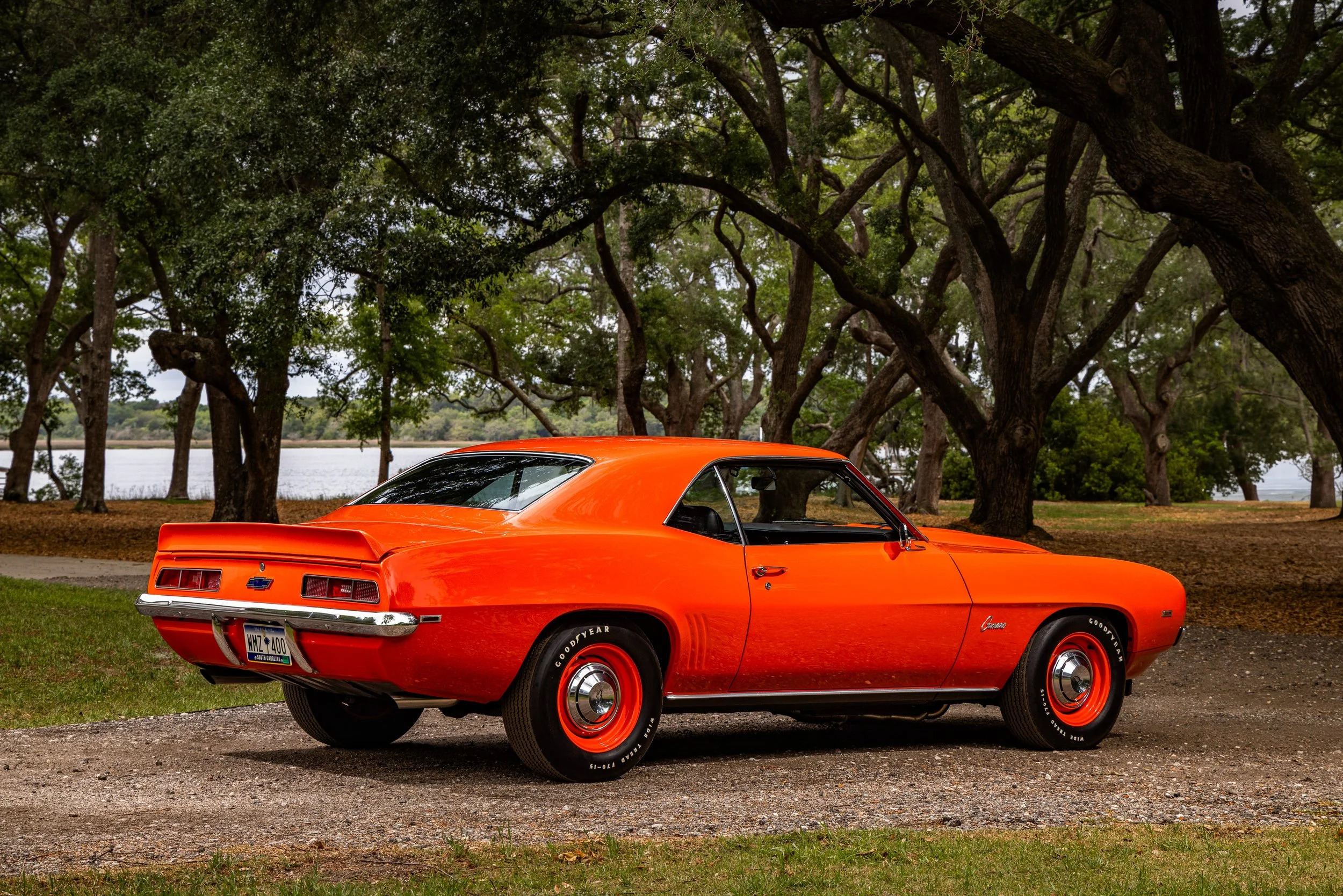 A classic orange Chevrolet Camaro car parked on grass near trees and a body of water.