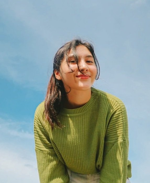 A young woman with dark hair, smiling at the camera, wearing a green sweater against a bright blue sky.