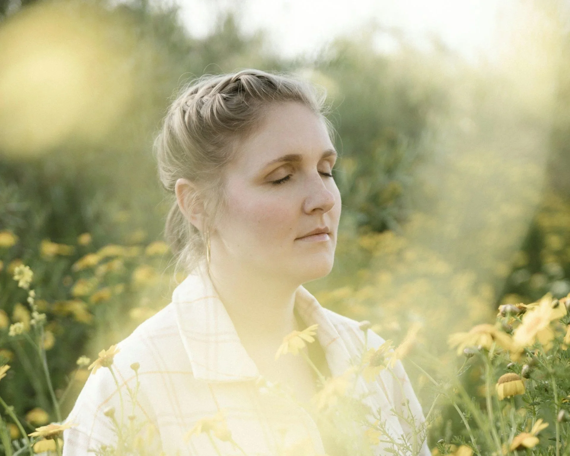 A woman with blonde hair styled in a braid, with her eyes closed, standing outdoors amidst yellow flowers and green foliage, wearing a light-colored outfit.