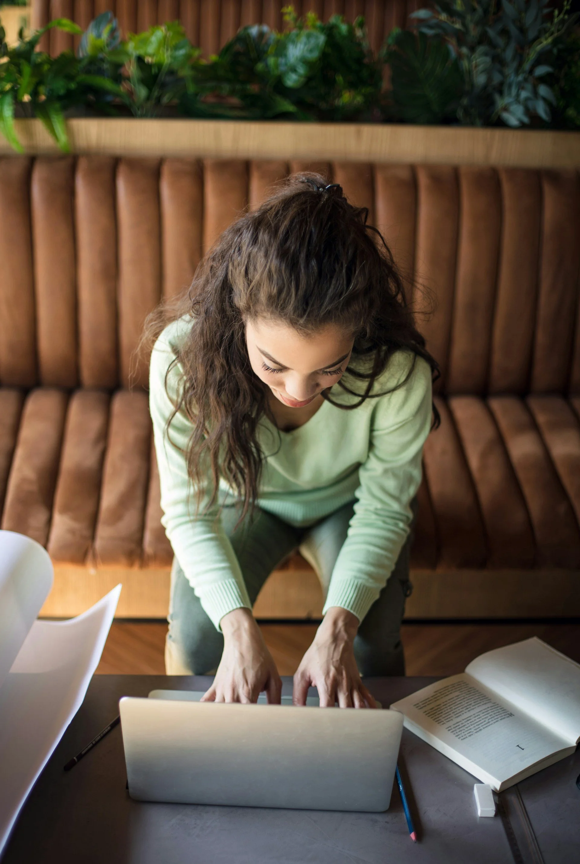 A woman with long, curly brown hair, wearing a light green sweater, sitting at a table, working on a laptop with open books, pens, and papers around her, positioned in front of a tufted brown leather bench with a wooden shelf filled with green plants behind her.