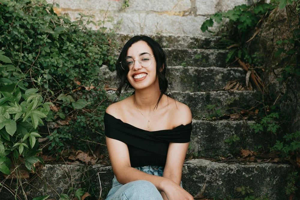 A young woman with black hair and glasses smiling and sitting on stone steps surrounded by green plants.
