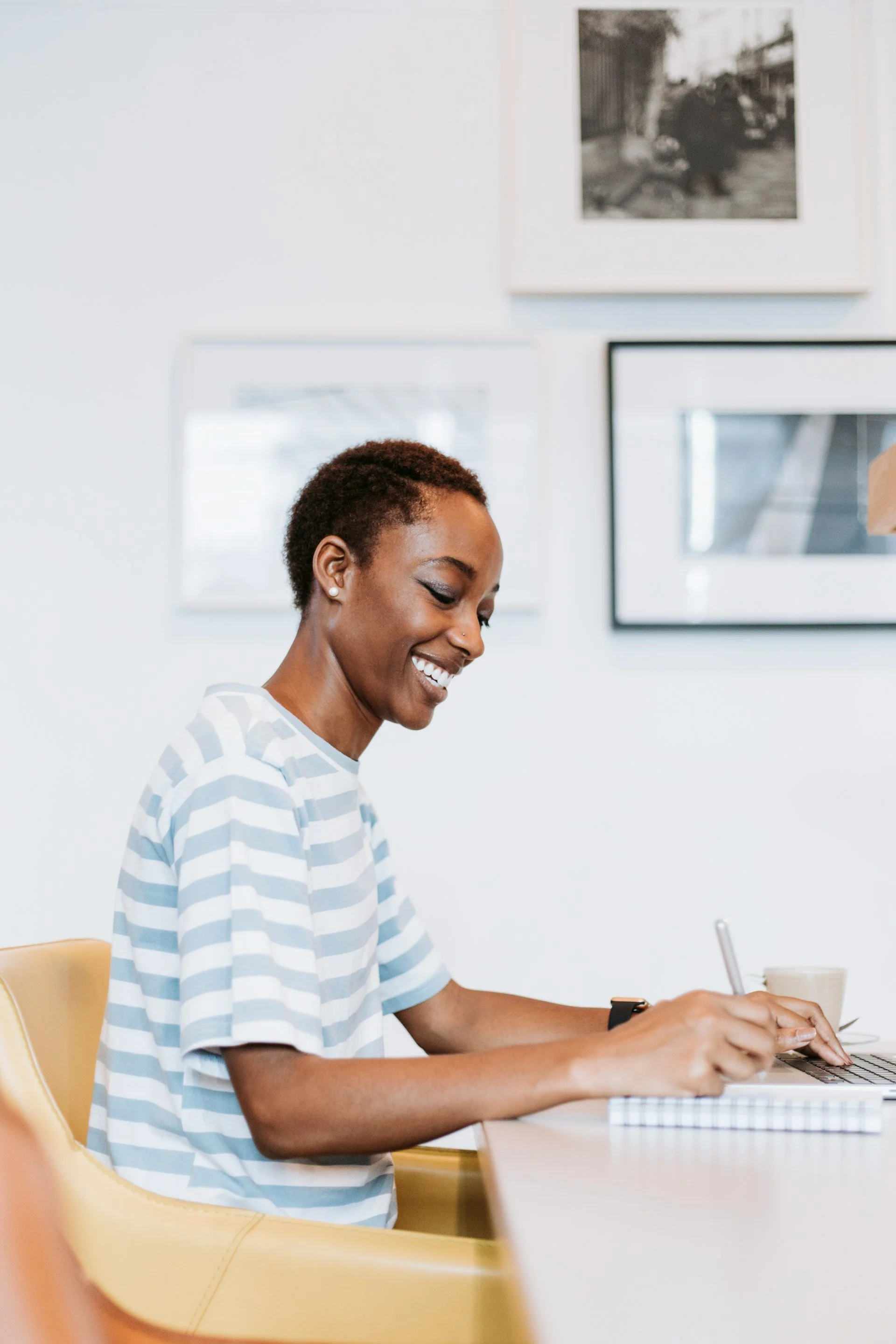 A woman with short hair, wearing a striped shirt, smiling and working on a laptop at a desk in a bright room with framed photos on the wall.