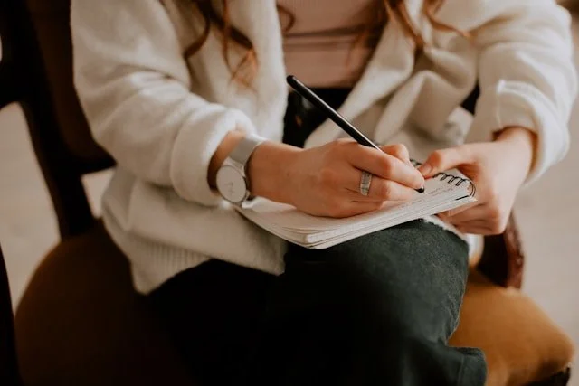 Person sitting and writing in a notebook with a pen, wearing a white hoodie and a watch.