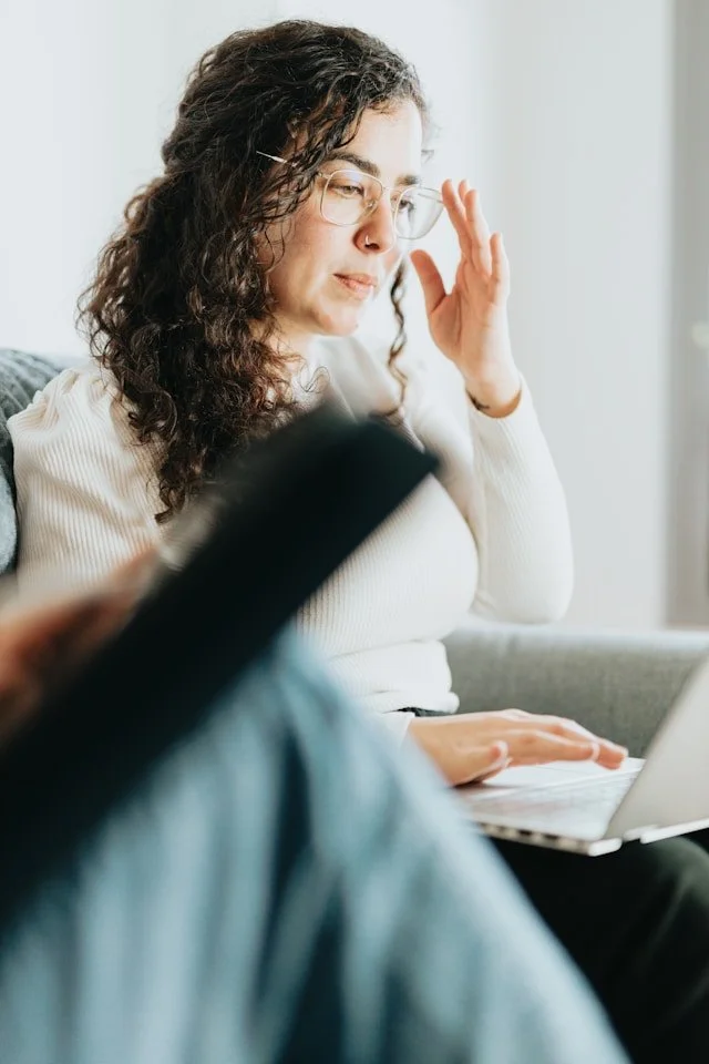 A young woman with curly hair and glasses adjusting her glasses while sitting on a couch with a laptop on her lap, in a well-lit room.