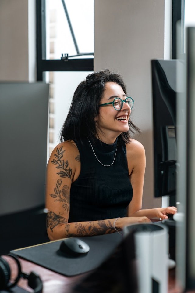 A woman with black hair, glasses, and tattoos on her arm is smiling and working at a computer in an office setting.