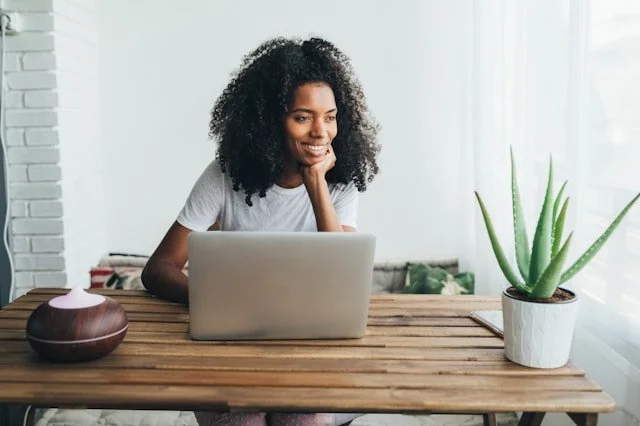 Young woman with curly hair working on a laptop at a wooden desk, smiling, in a bright room with a plant and decorative items.