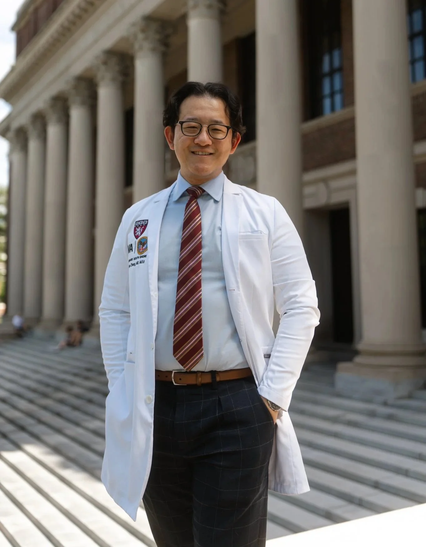 A smiling man in a white medical coat and striped tie stands on steps in front of a historic building with columns and large windows.