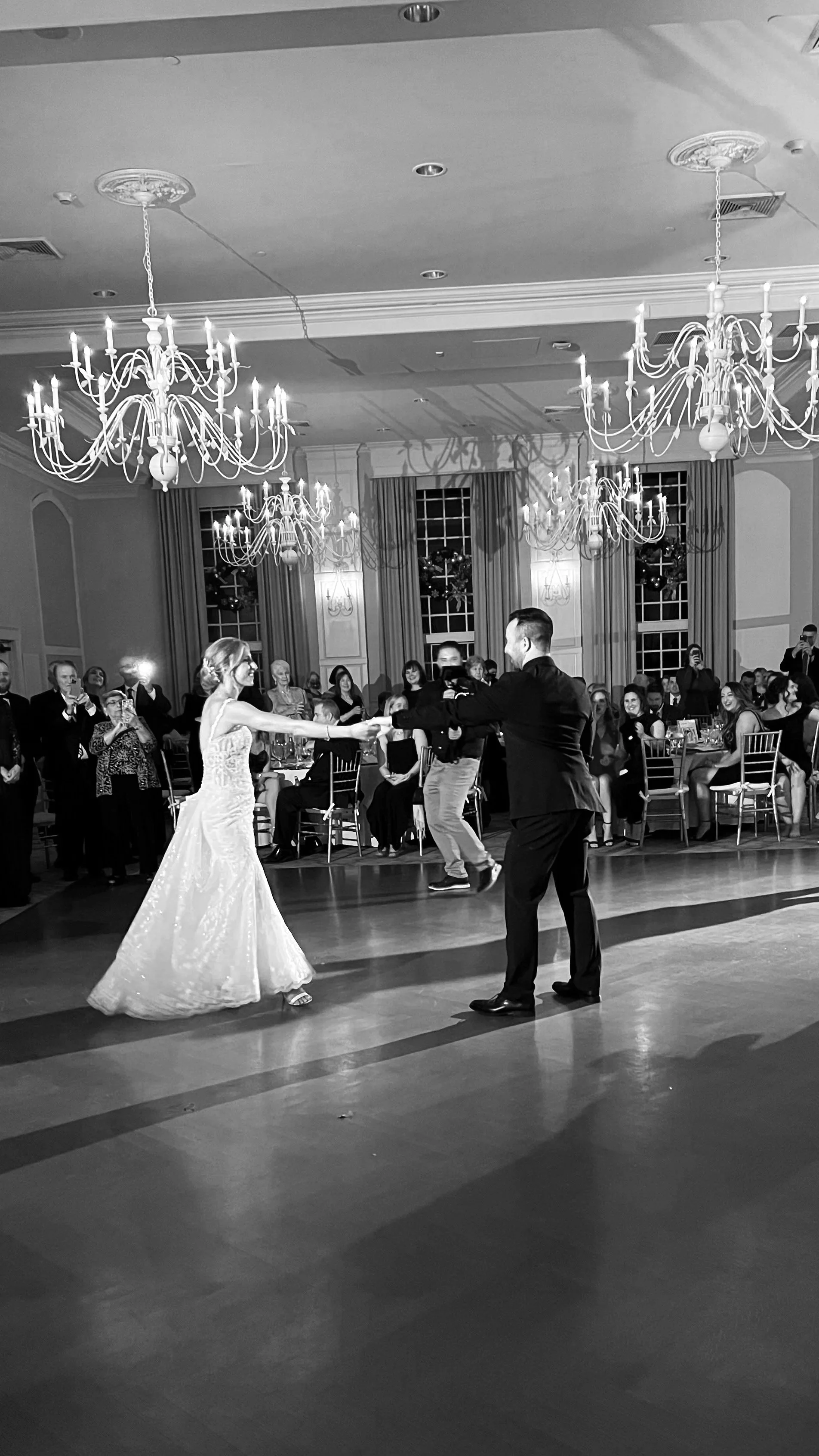 A bride and groom dancing at their wedding reception with guests watching and taking photos in a decorated ballroom with chandeliers.