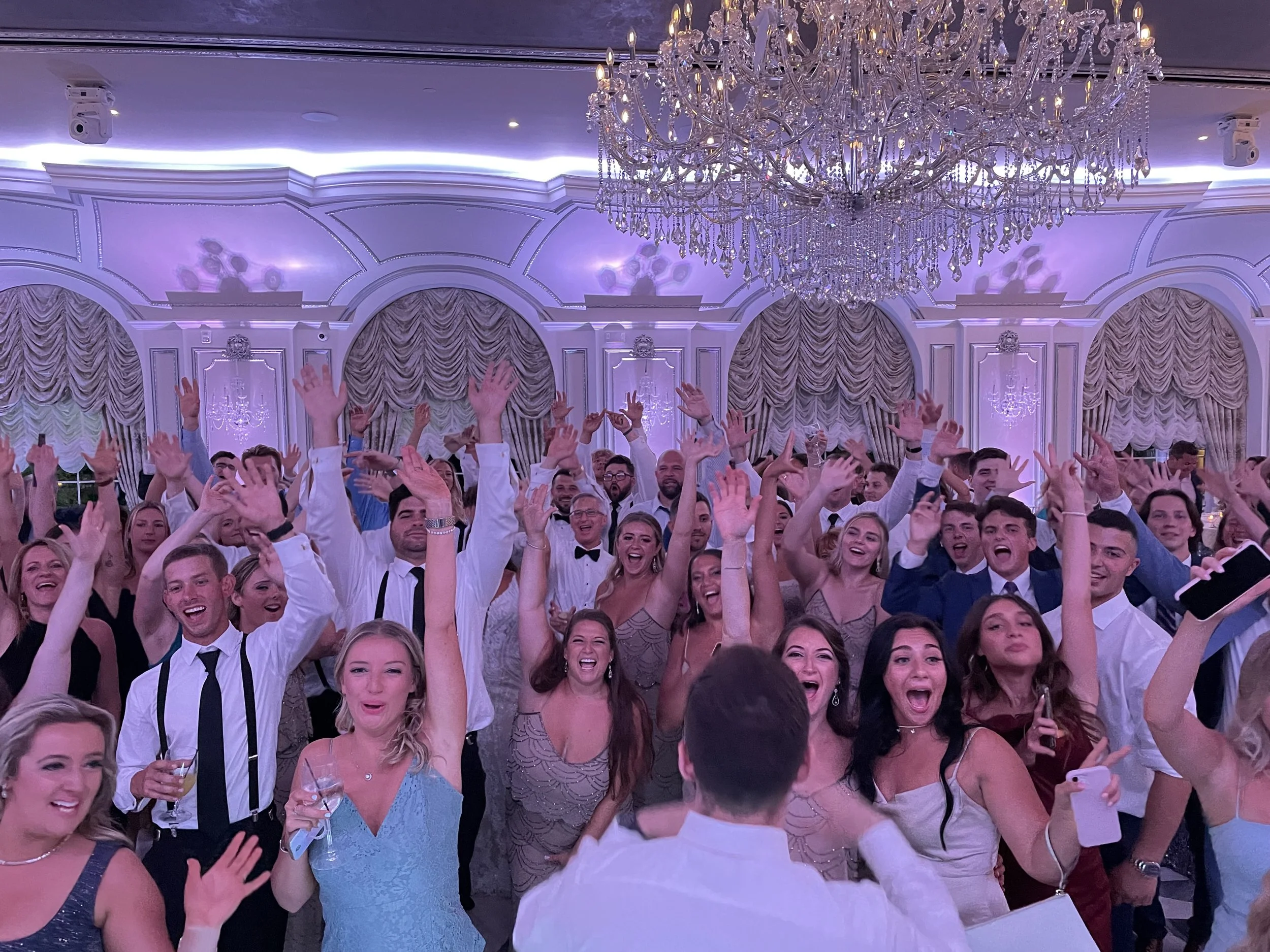 A large crowd of people at a formal event, all dressed in elegant attire, raising their hands and smiling under a grand chandelier in a luxurious decorated room.