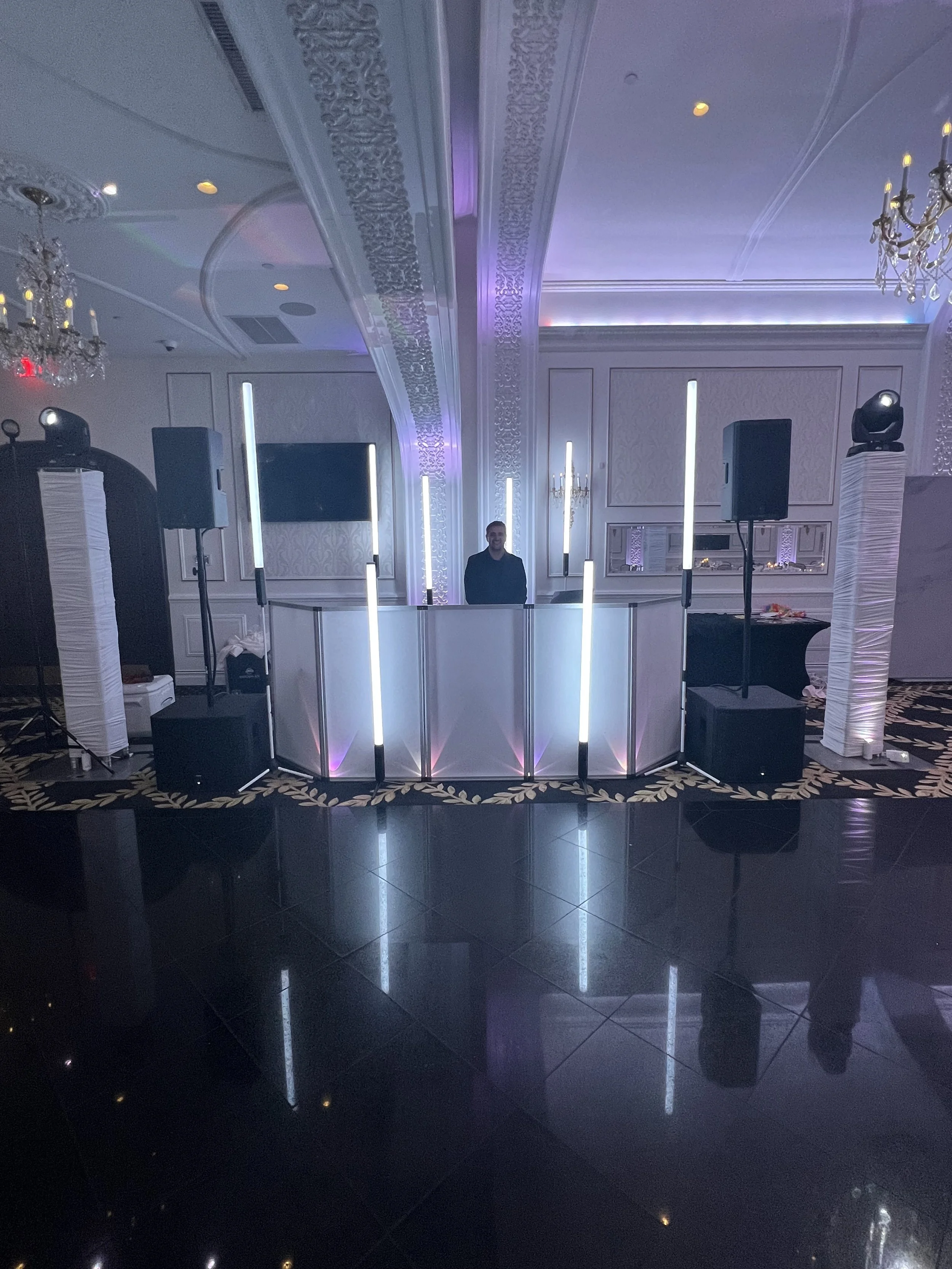A person standing behind a DJ booth in a decorated event hall with ornate ceiling moldings, chandeliers, and vertical light bars.