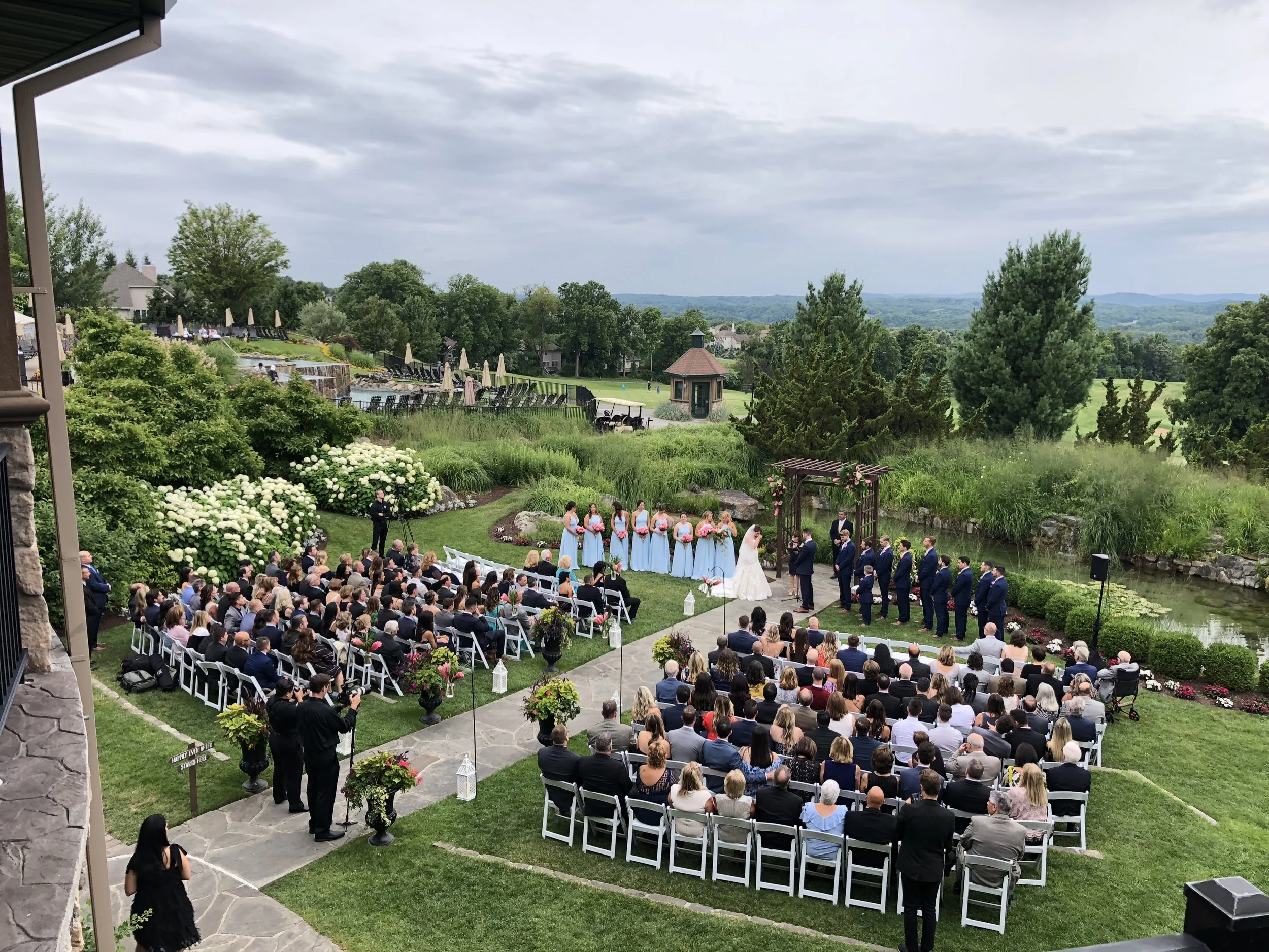 Outdoor wedding ceremony with guests seated on white chairs, bridal party standing under a wooden arch, surrounded by greenery, a pond, and scenic mountains in the background on a cloudy day.
