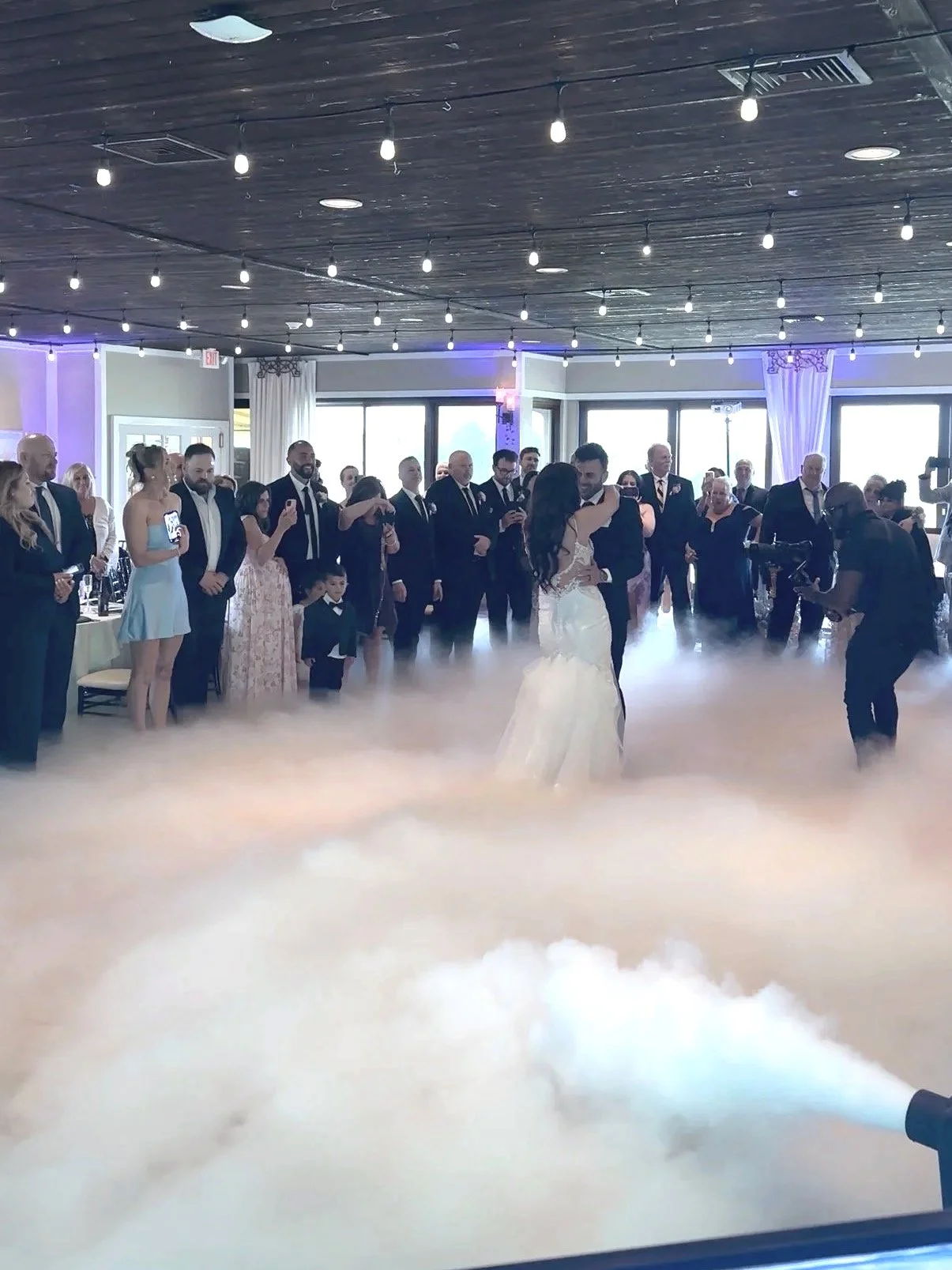Bride and groom dancing in a fog-filled dance floor at their wedding reception, surrounded by guests watching and taking photos in a decorated indoor venue with string lights.