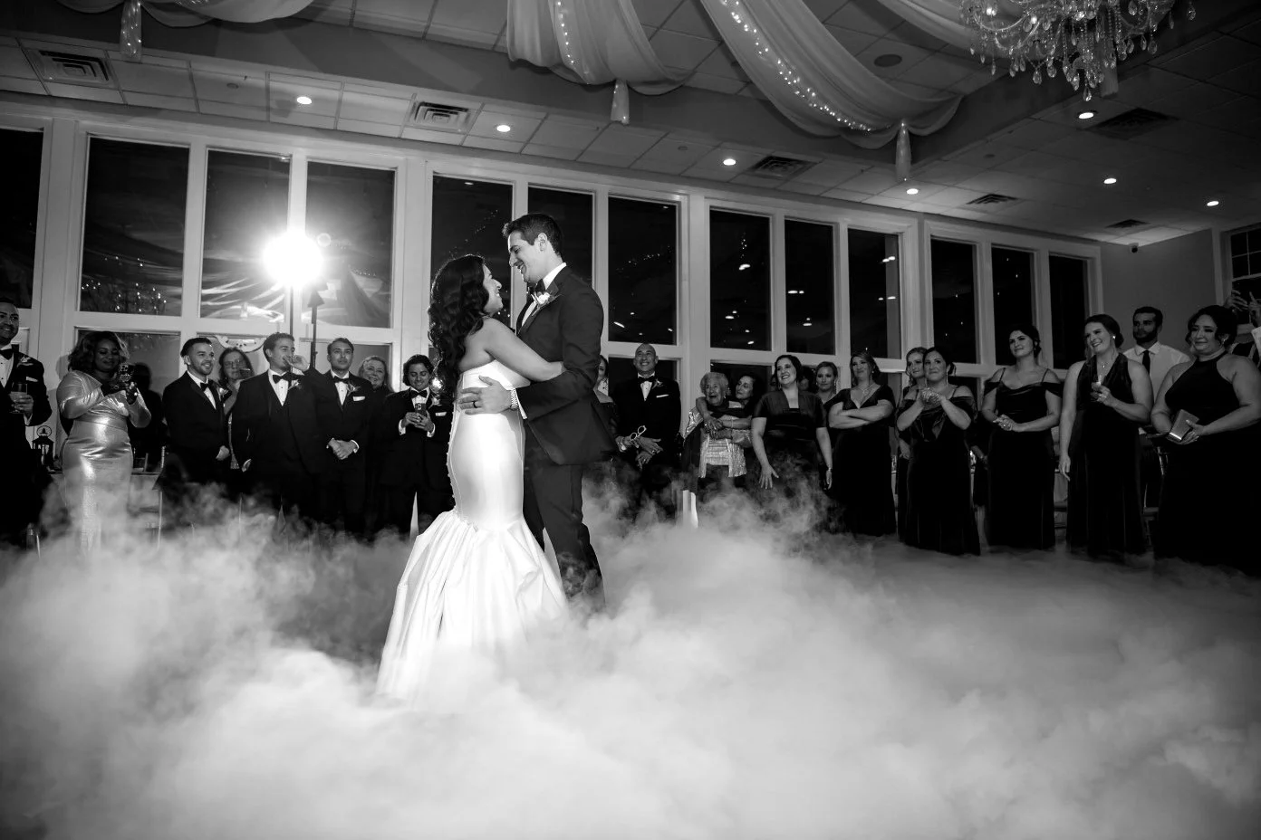 A bride and groom dance in the center of a wedding reception surrounded by friends and family, with fog on the floor and a large window behind them.