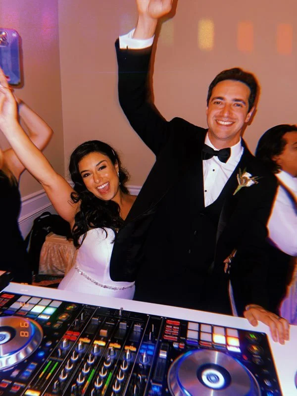 A bride and groom celebrating at a wedding reception, dancing with smiles and raised arms, with a DJ deck in the foreground.