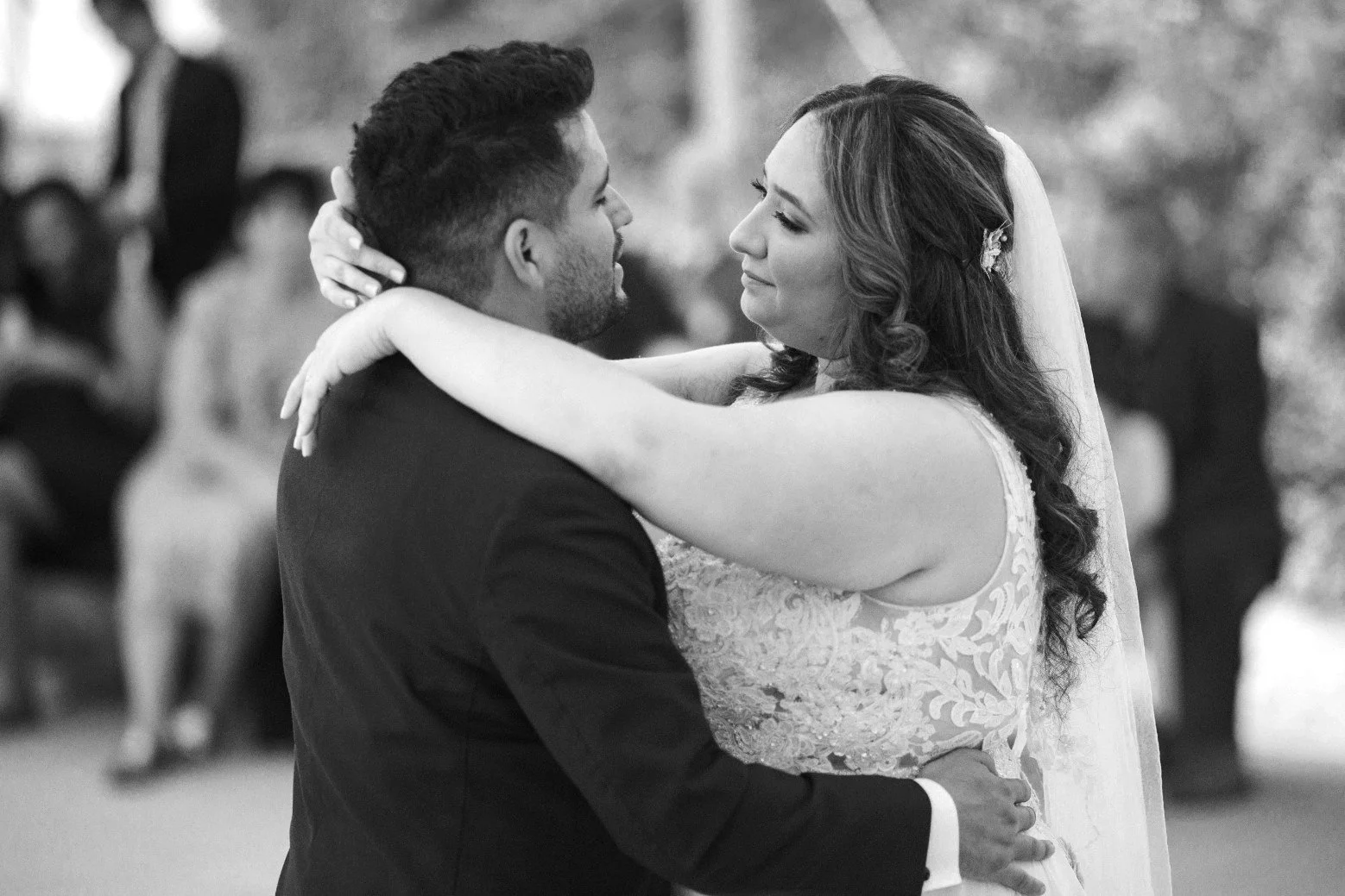 A black and white photo of a bride and groom sharing their first dance at their wedding. The bride has long, wavy hair and a lace wedding dress, and the groom is dressed in a suit. They are gazing into each other's eyes, embracing closely.