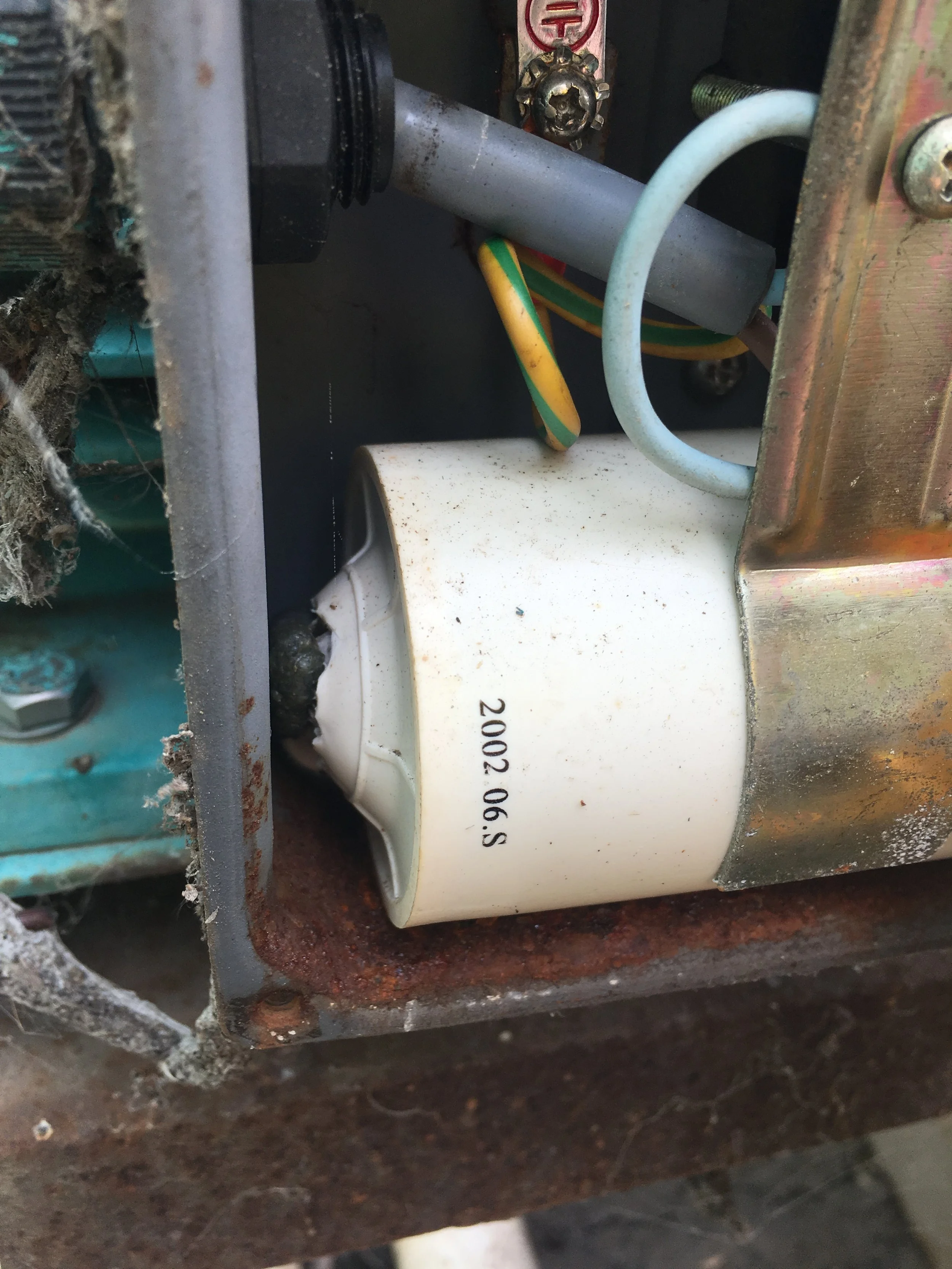 Close-up of an electrical component, showing a white cylindrical capacitor with printed markings, mounted on a rusty metal surface with some wires and other electrical parts surrounding it.