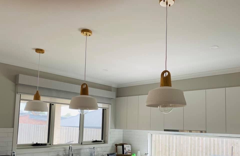 Three white hanging pendant lights with wooden accents over a kitchen counter with white cabinets and windows.