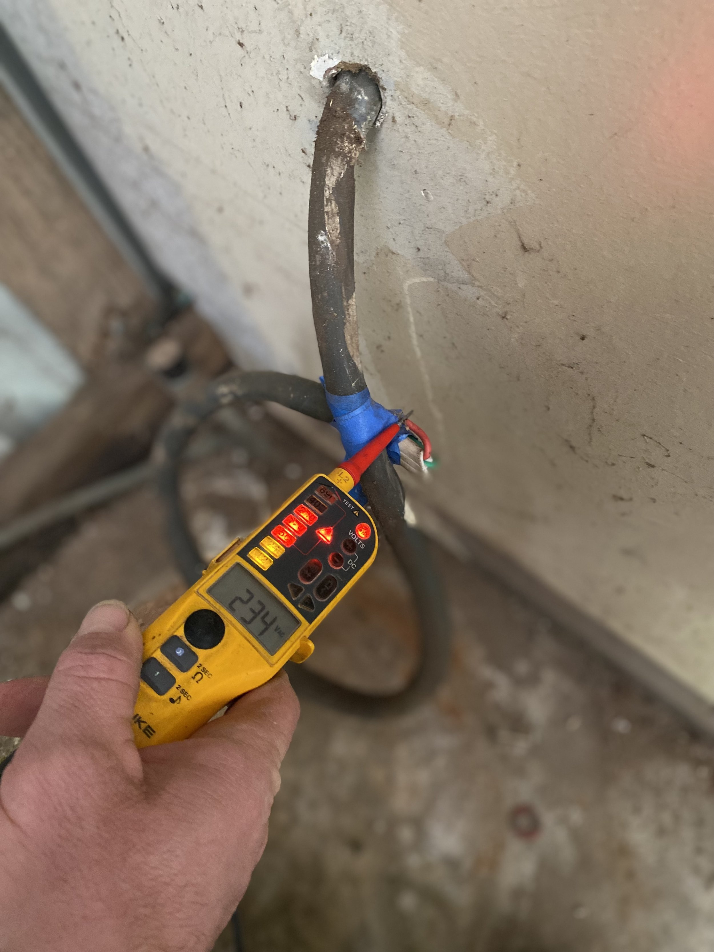 A person holding a yellow digital multimeter measuring 234 volts, with wires connected to a wall electrical outlet.