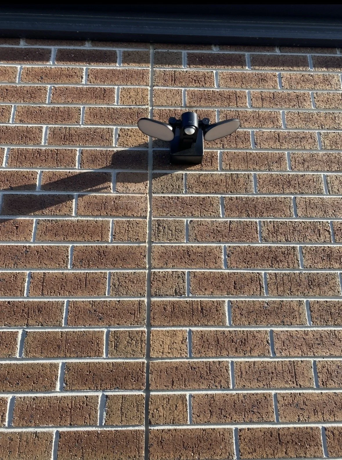 A black outdoor security light attached to a brick wall with a shadow cast below it.