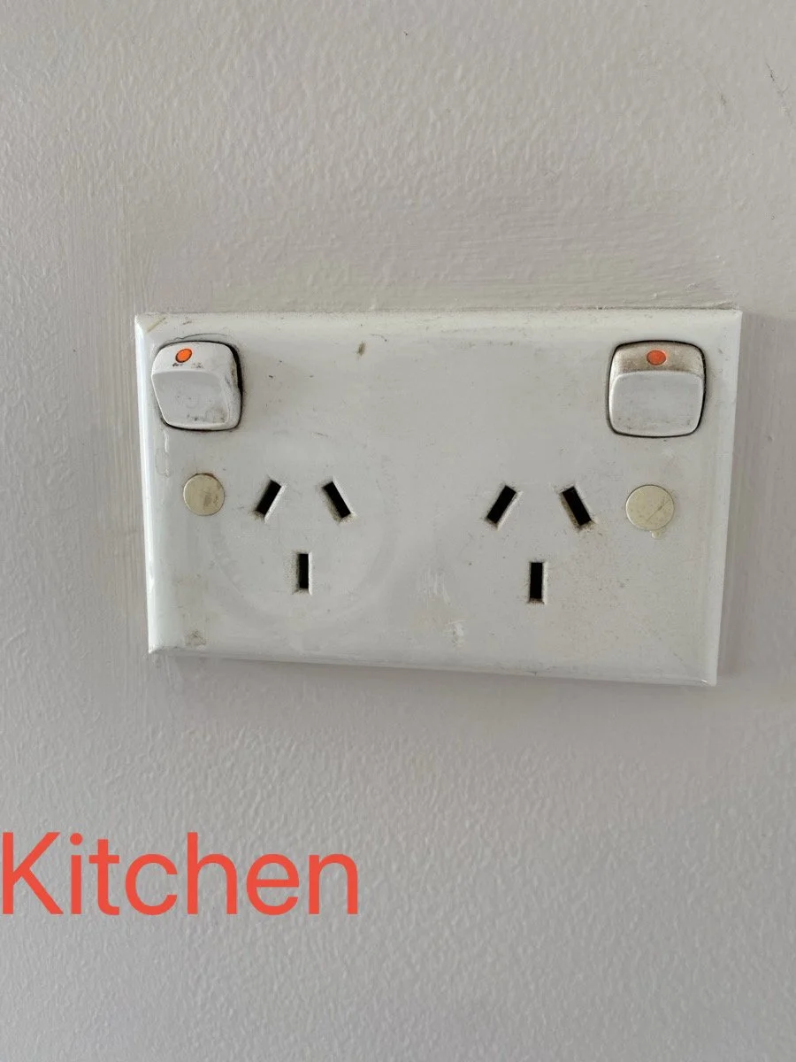 A white kitchen electrical outlet with two switches, two power sockets, and a wall painted in light color. The switches have orange indicator lights, and the outlet shows some dirt and wear.