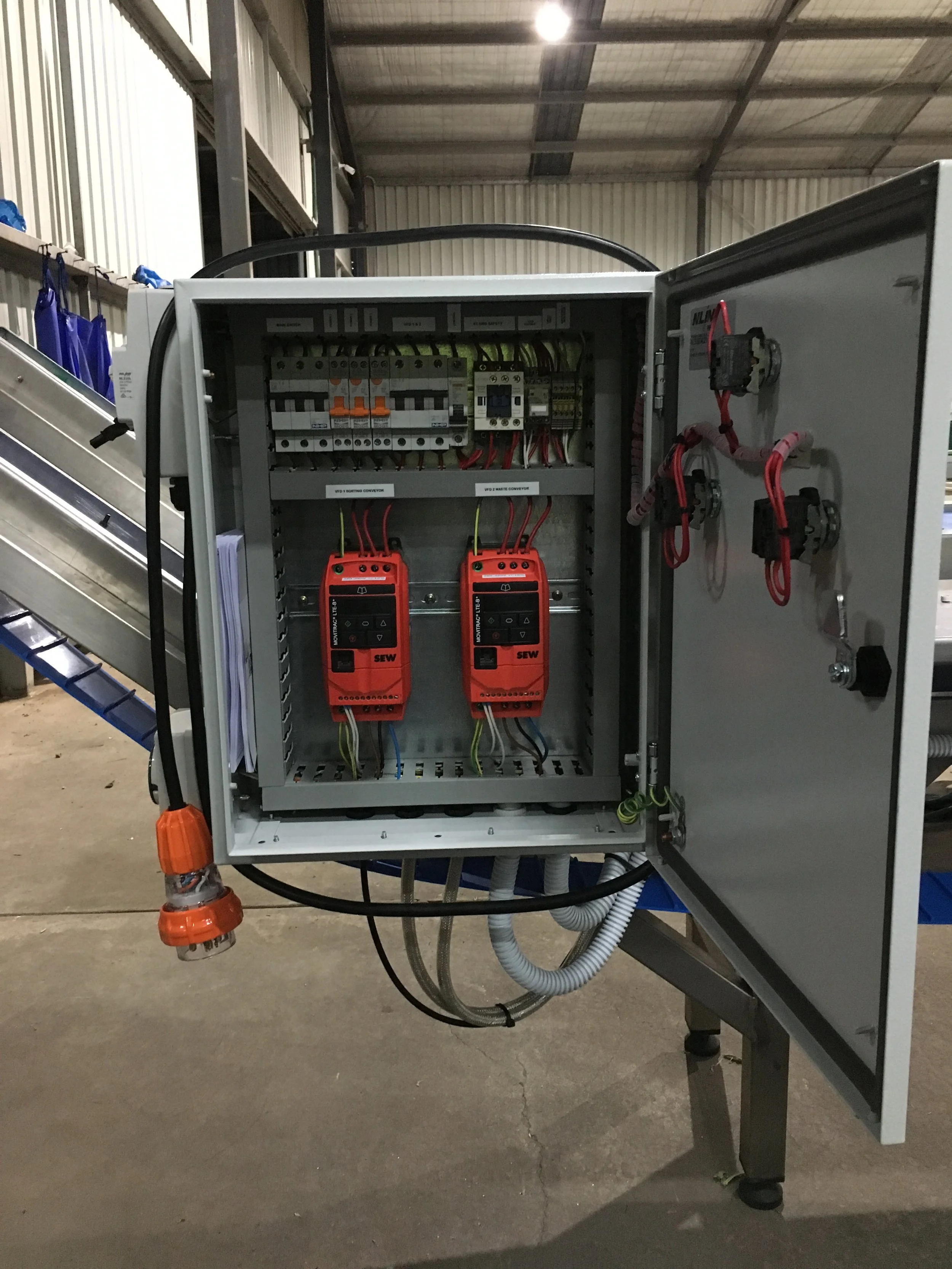 Electrical control panel with wiring and measuring devices inside a metal enclosure, located in a warehouse or industrial setting.
