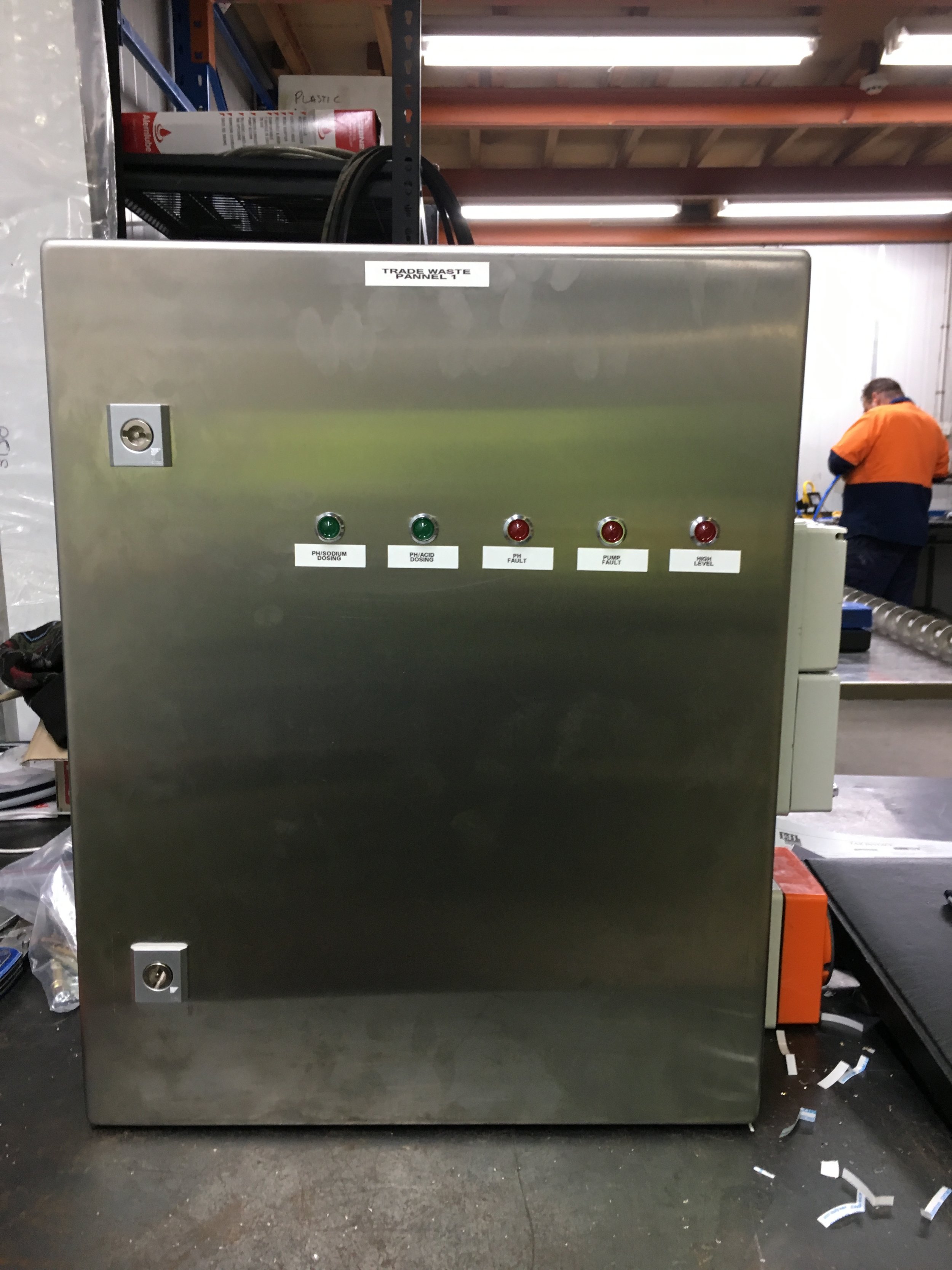 Industrial metal cabinet with labeled warning lights and a keypad, placed in a warehouse with storage shelves and a worker in the background.